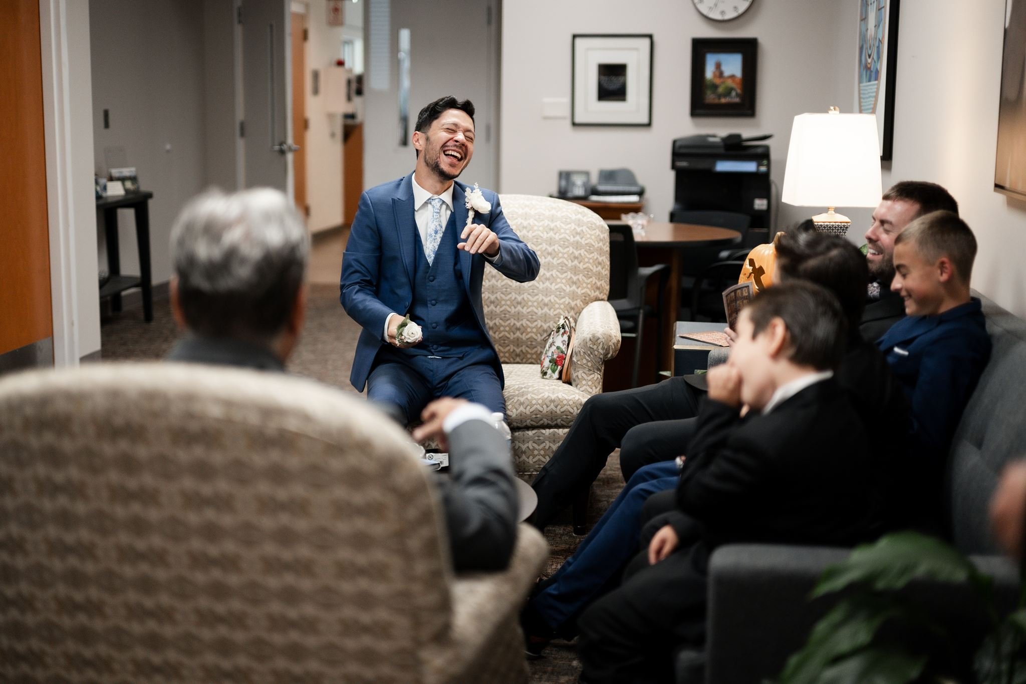 A man in a blue suit laughing and sitting in a chair surrounded by people in suits in a room decorated with framed pictures on the wall before his wedding at St. Mathews Catholic Church in Charlotte, NC.
