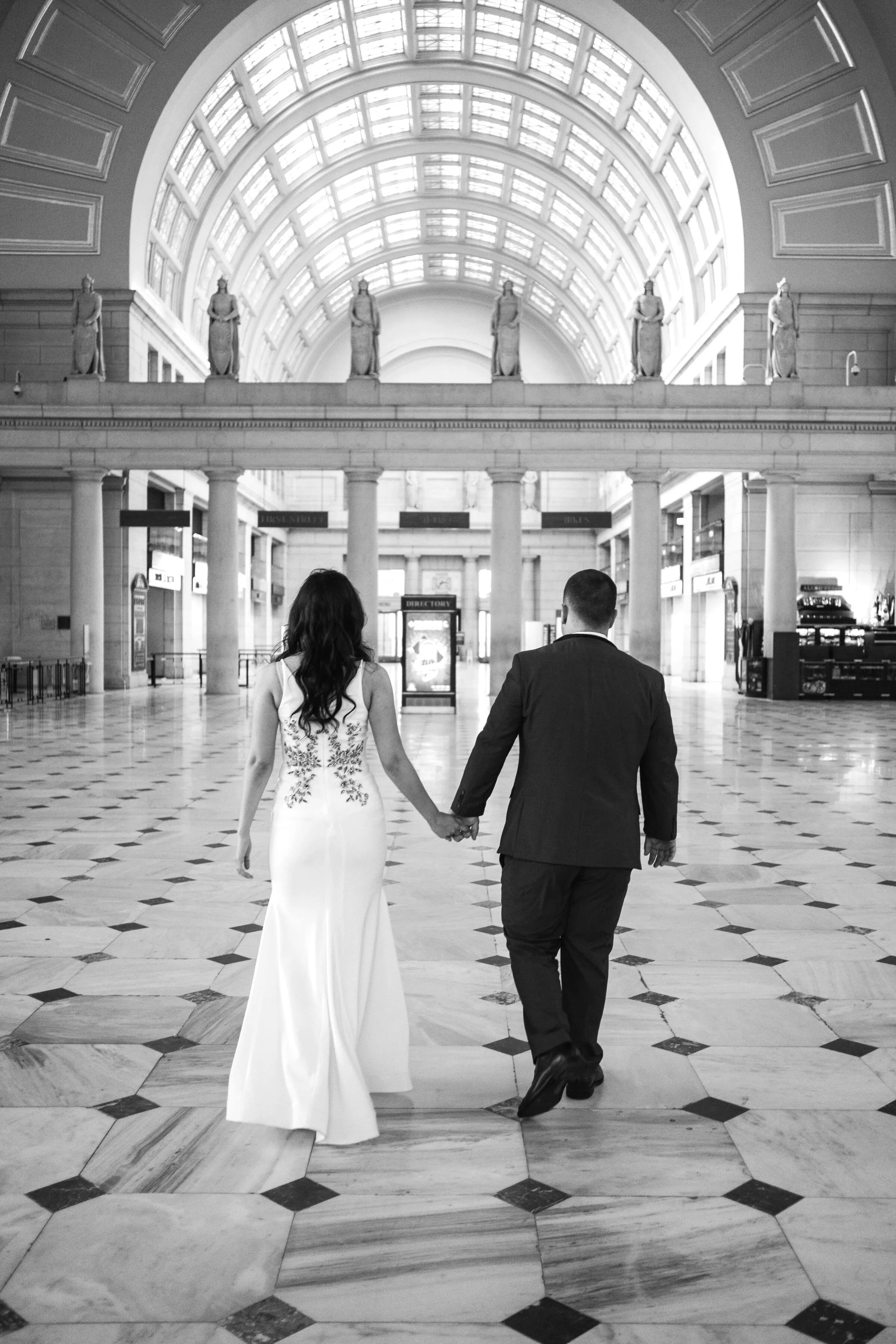 A bride and groom walking hand in hand in a grand indoor space with high arched glass ceilings, statues, and a patterned marble floor in DC.
