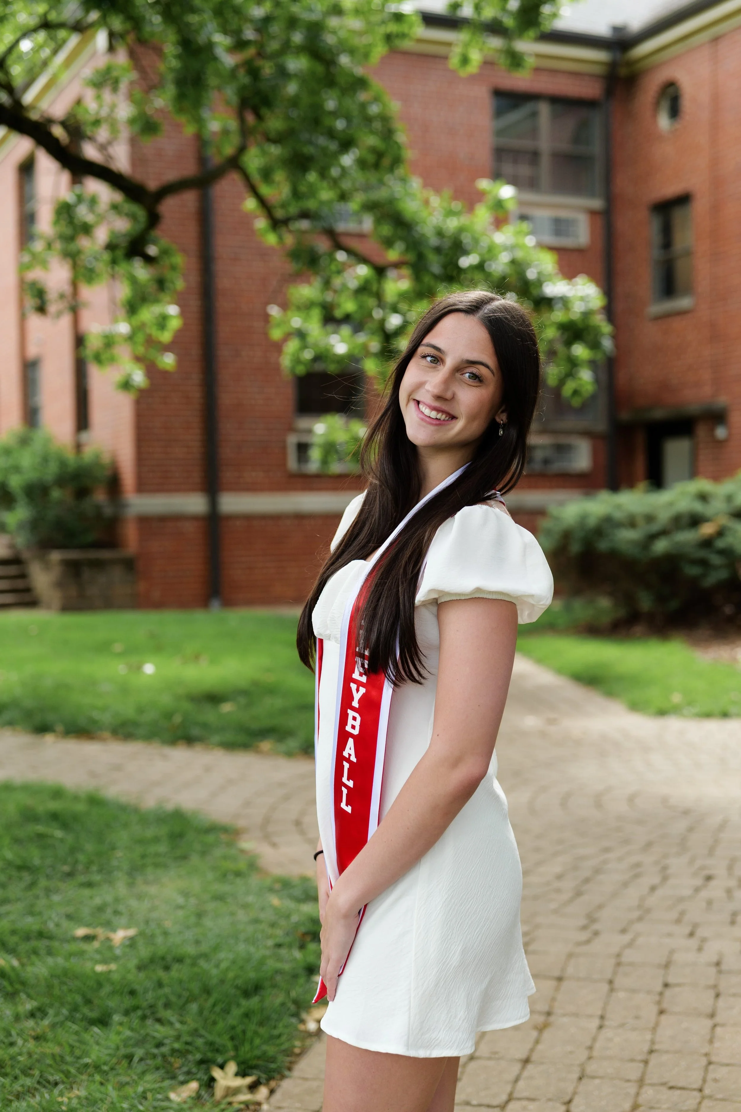 A young woman with long dark hair smiling at camera outdoors, wearing a white dress with a red and white ribbon during a graduation photo session in Pineville, NC