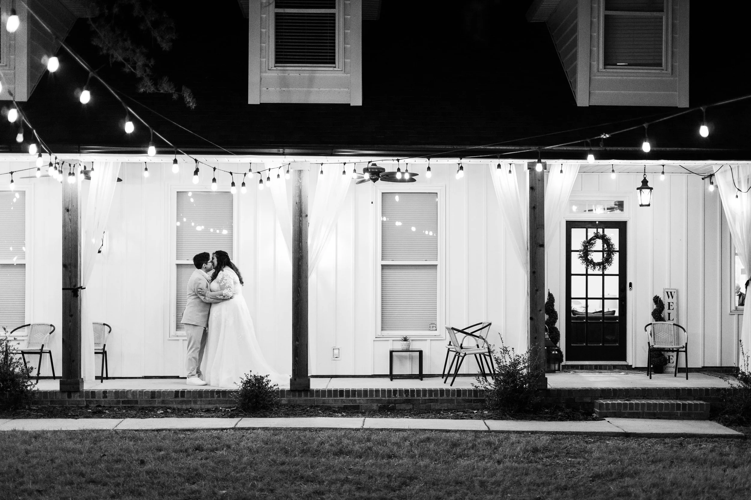 appears to be a black and white wedding photo from Brandon Pickett Photography of two gay married woman kissing on a porch at night.