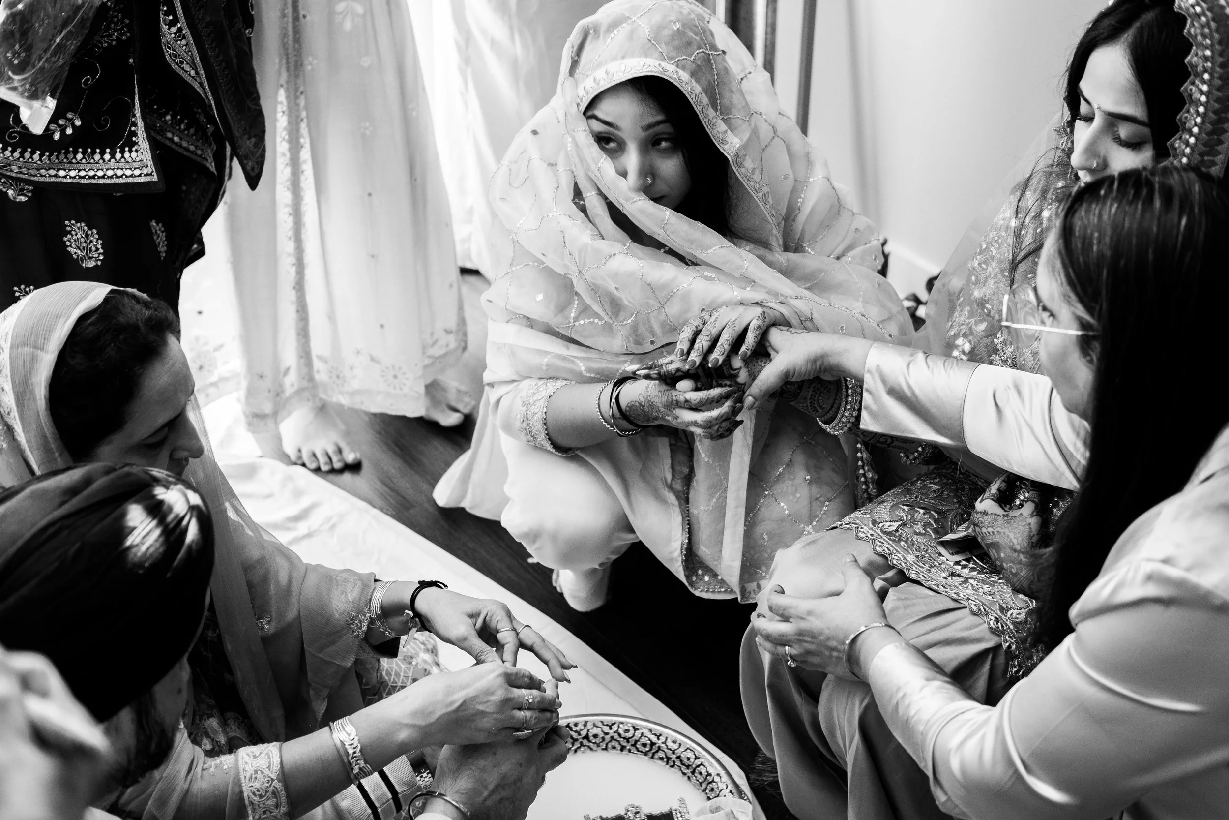 A group of women dressed in traditional South Asian attire, involved in a ceremonial or religious activity, with one woman in the center wearing a dupatta over her head and others surrounding her, some holding her hands.