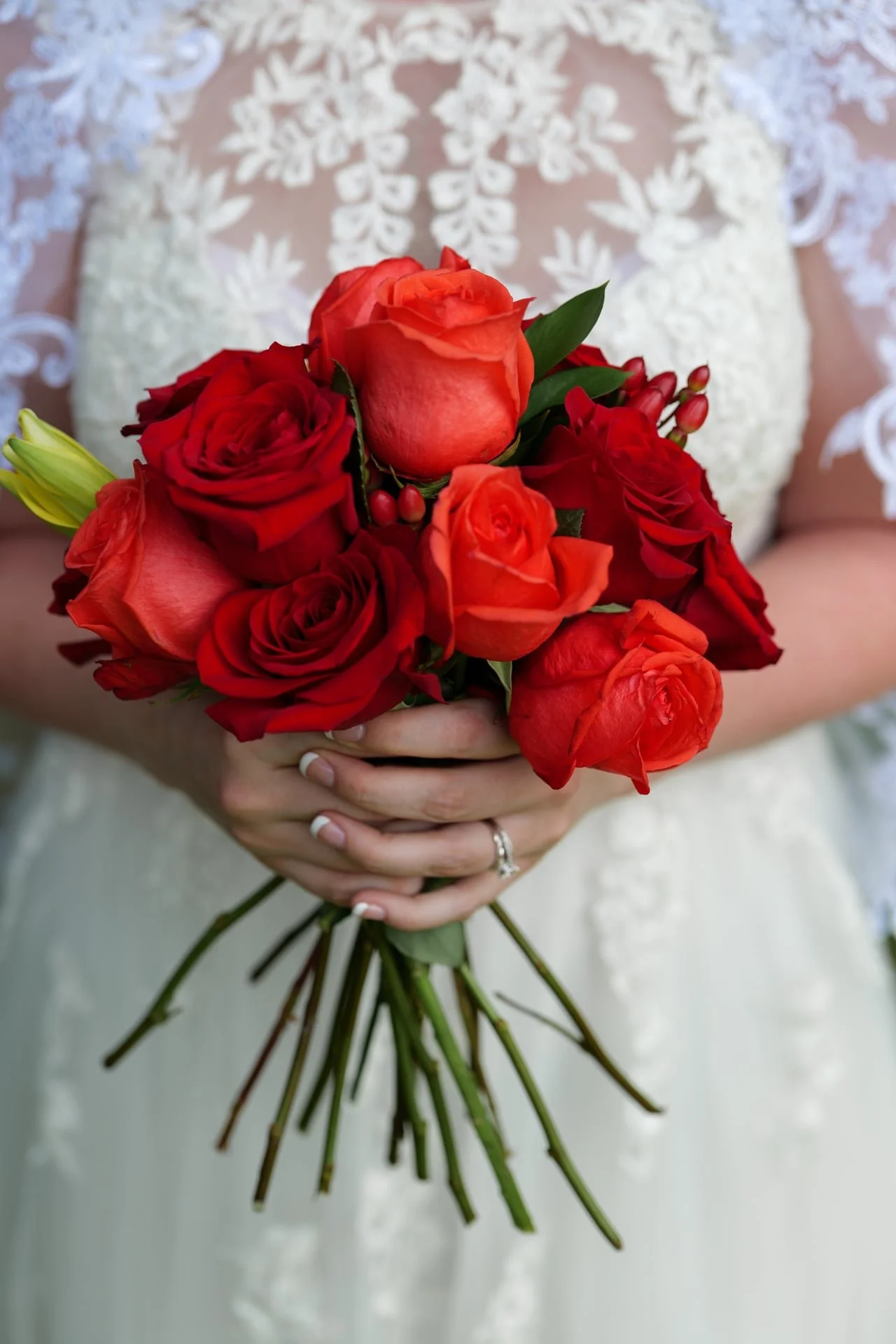 A bride holding a bouquet of red and orange roses with greenery in front of her lace wedding dress.