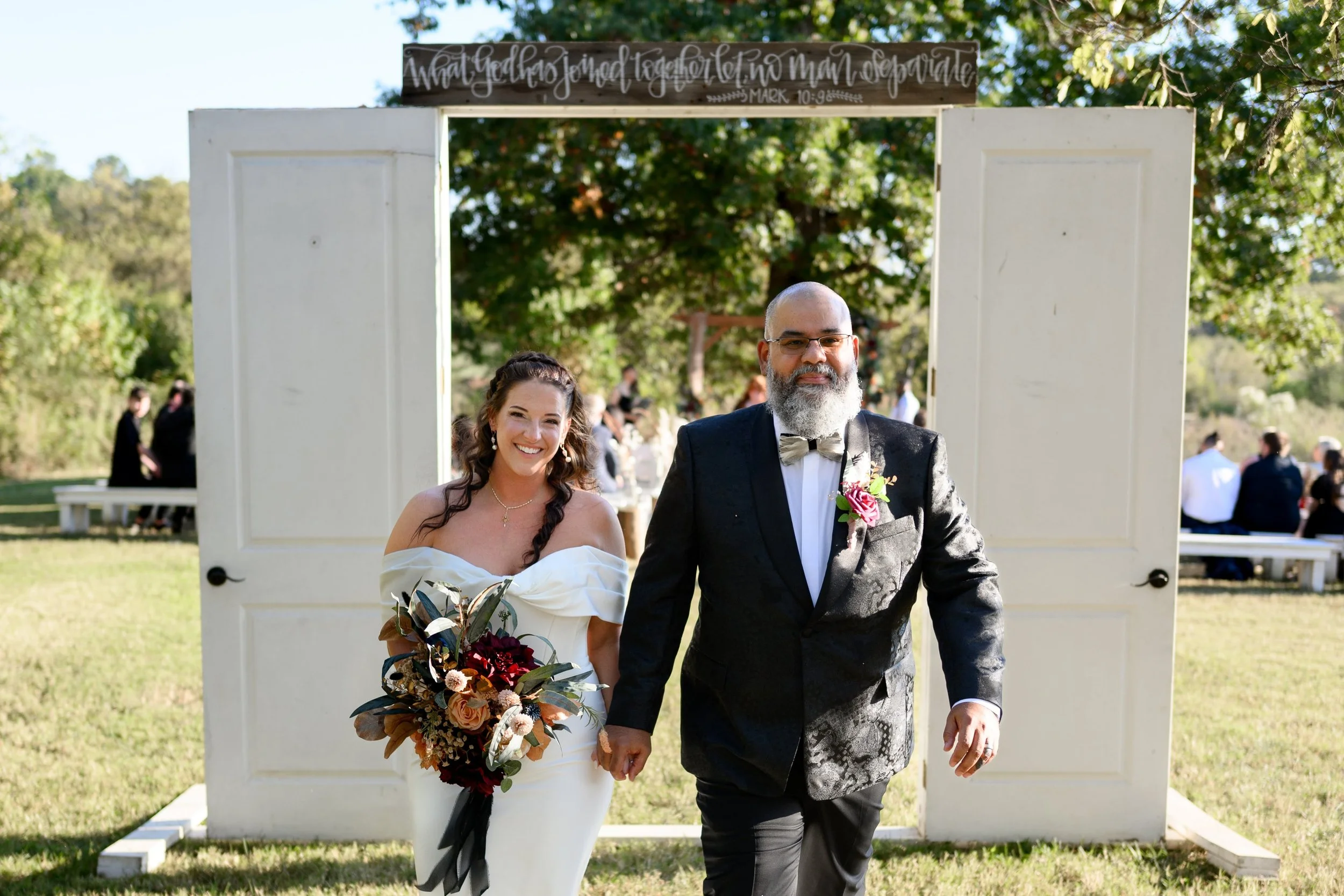 A bride and a man, possibly her father, walking outdoors after a wedding ceremony. The bride is wearing a white off-shoulder wedding dress and holding a bouquet of flowers, while the man is dressed in a black suit with a bow tie and boutonniere. They