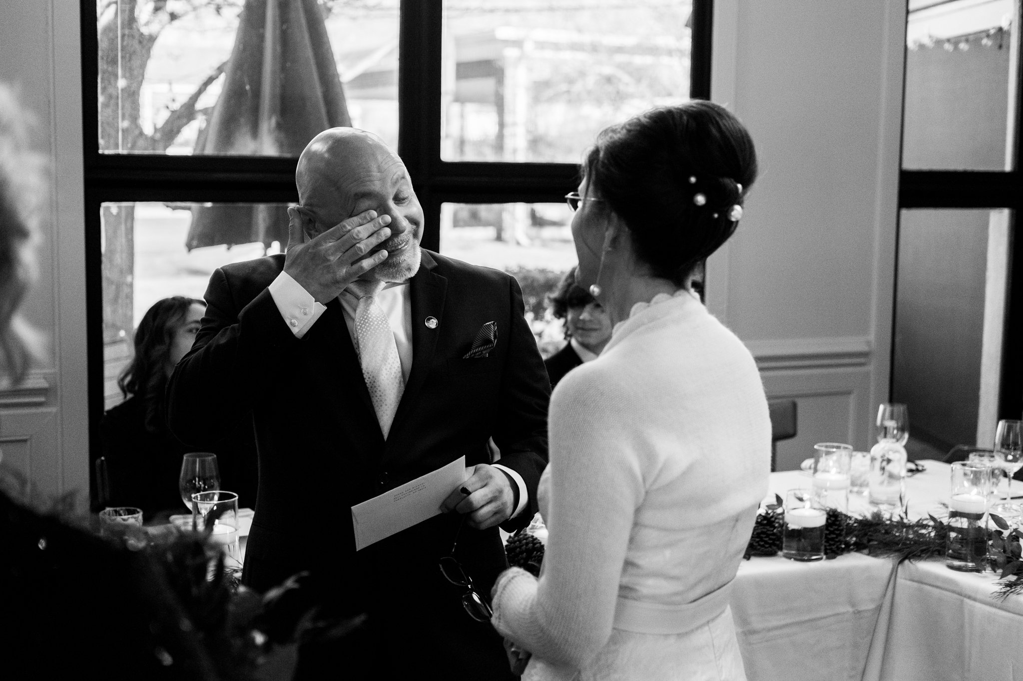 A man in a suit appears to be giving a heartfelt speech while wiping away tears during a wedding reception; a woman in a white dress, likely the bride, listens attentively.