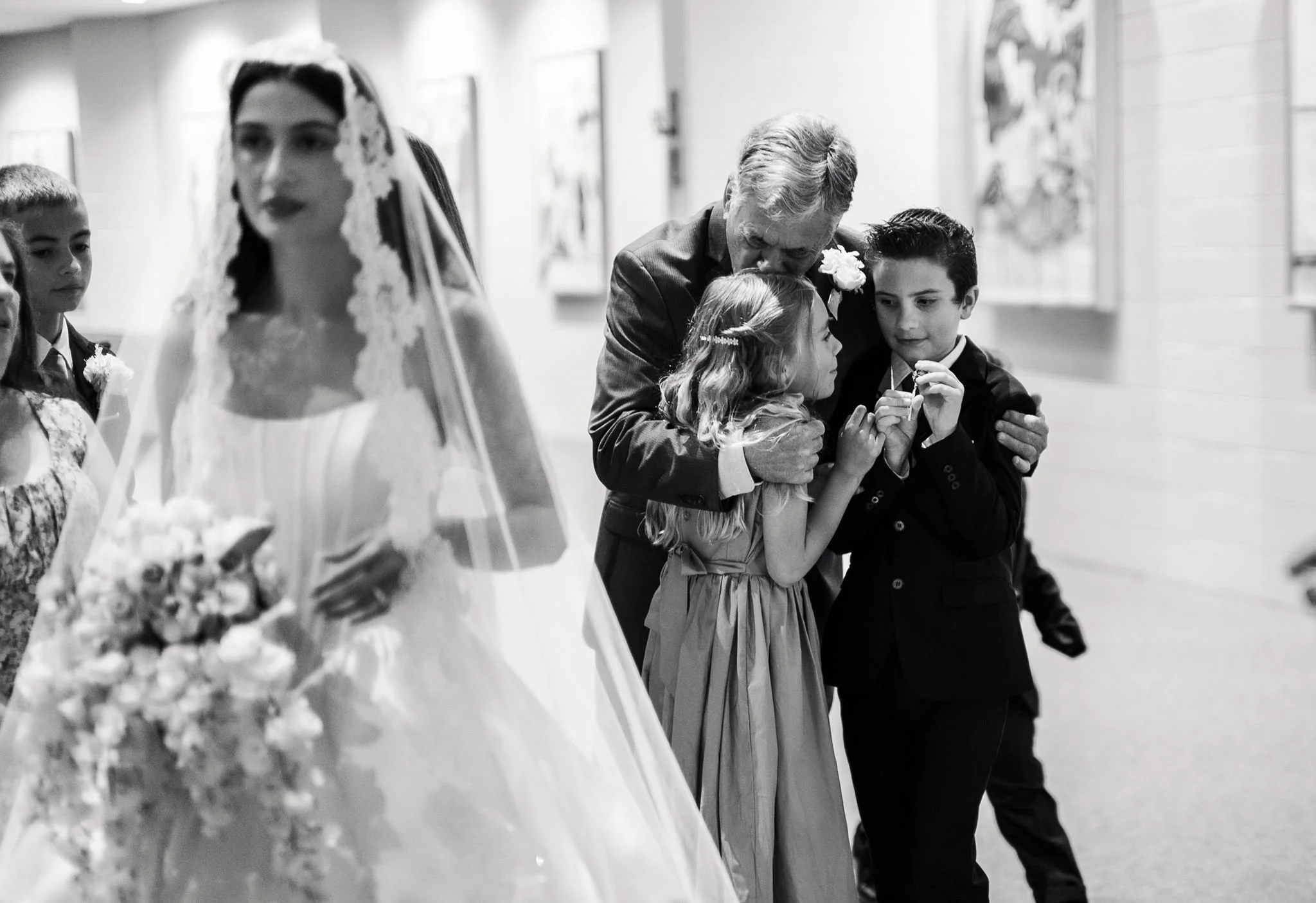 A black and white photograph of a wedding ceremony where a man and a girl are sharing a moment of prayer or solemn reflection, surrounded by children in formal attire. The girl has a dress and a hair accessory, while the man is wearing a suit, and they are close together with their heads bowed.