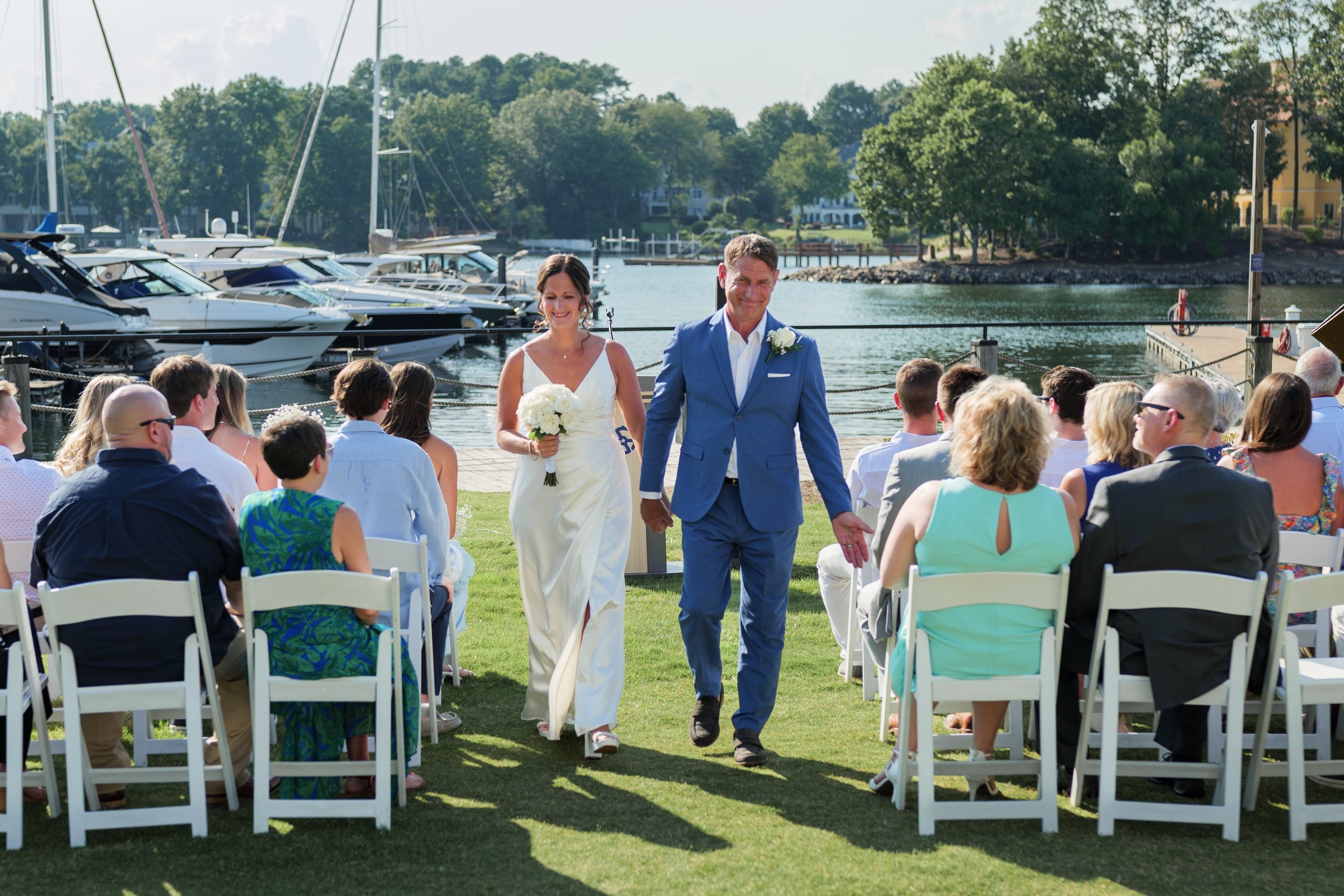 A newlywed couple walking down the aisle at an outdoor wedding ceremony near a marina with boats, with guests seated on white chairs on a grassy area on a sunny day.
