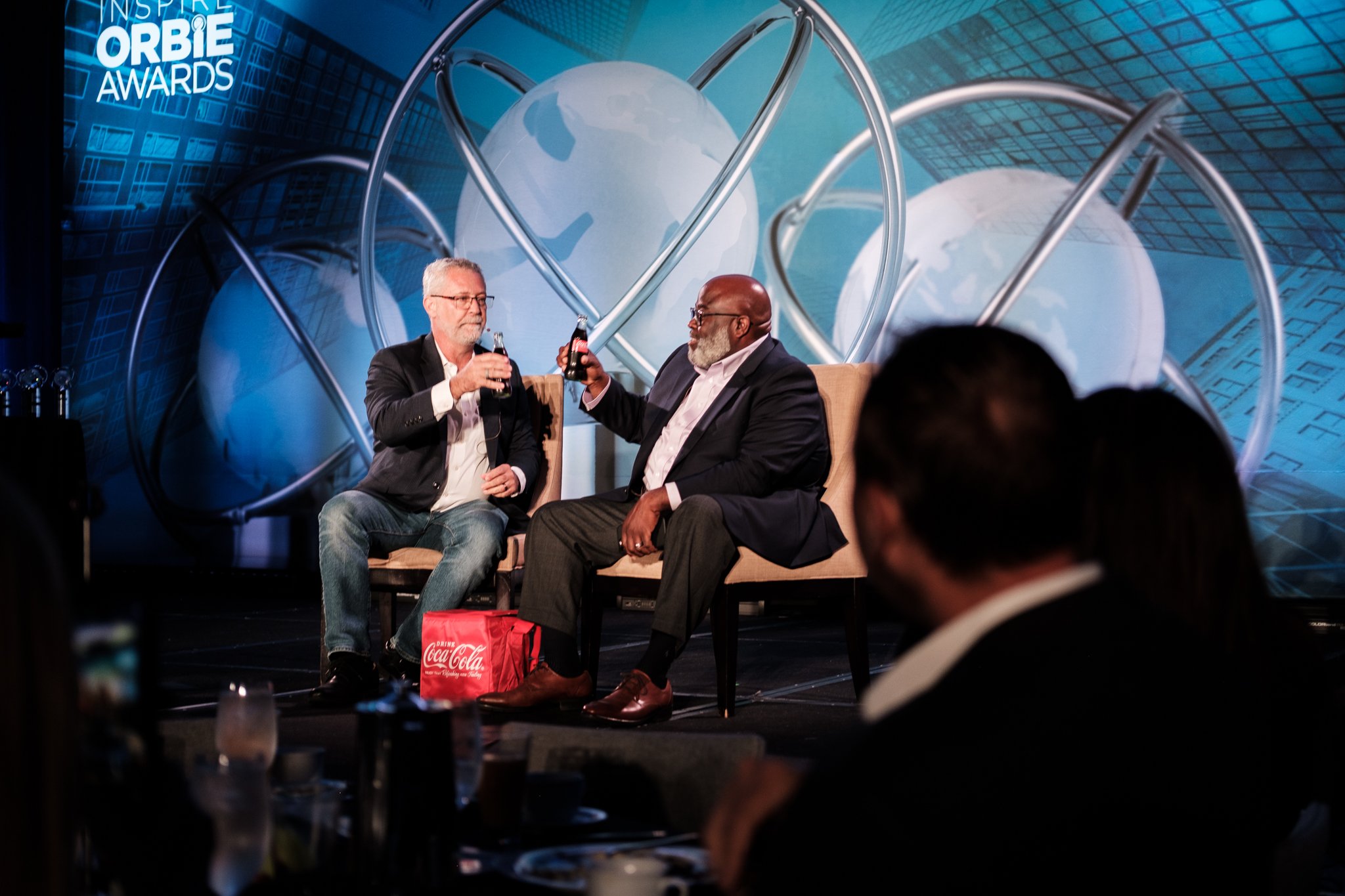Two men sitting on stage during an award show, clinking bottles of Coca-Cola. The background features large digital graphics with globes and orbit-like rings, and the words 'Inspire Orbie Awards'.