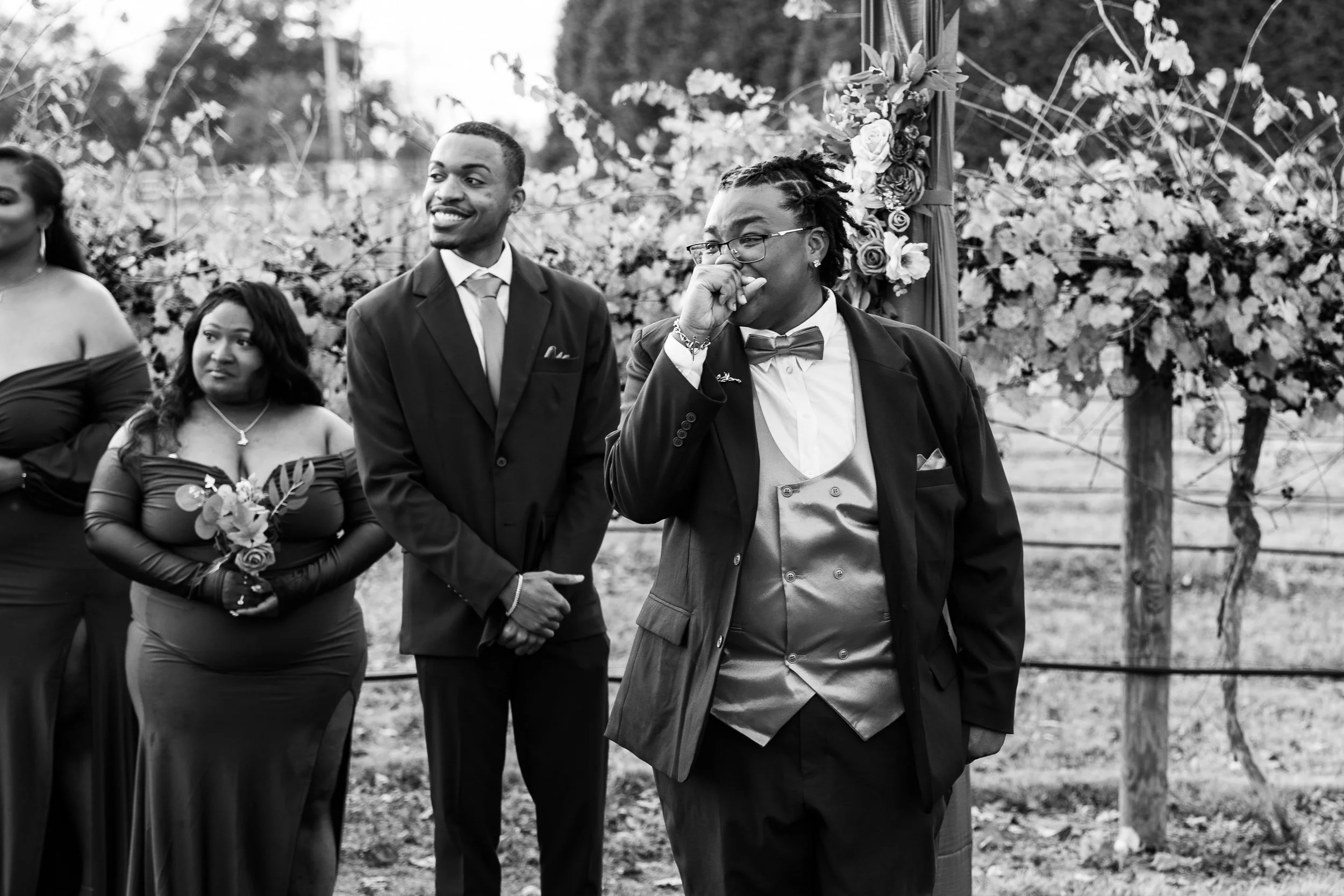 A black-and-white photo of a wedding ceremony outdoors, featuring a group of people standing in front of an arch decorated with flowers. One woman is holding a bouquet, and a man is wearing a tuxedo. The atmosphere is joyful and celebratory.