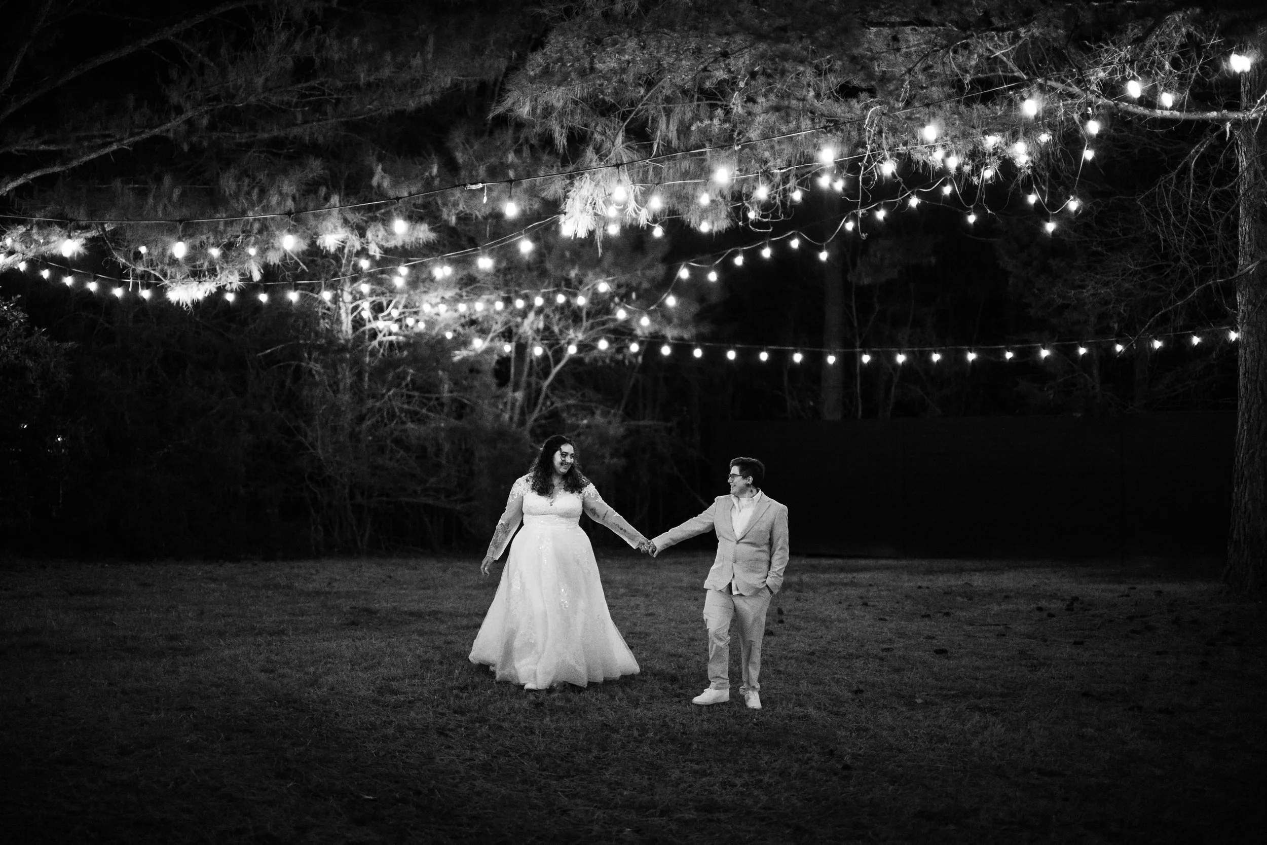 Black and white outdoor wedding reception at night with string lights overhead, a bride and groom holding hands and walking in a field, trees in the background.