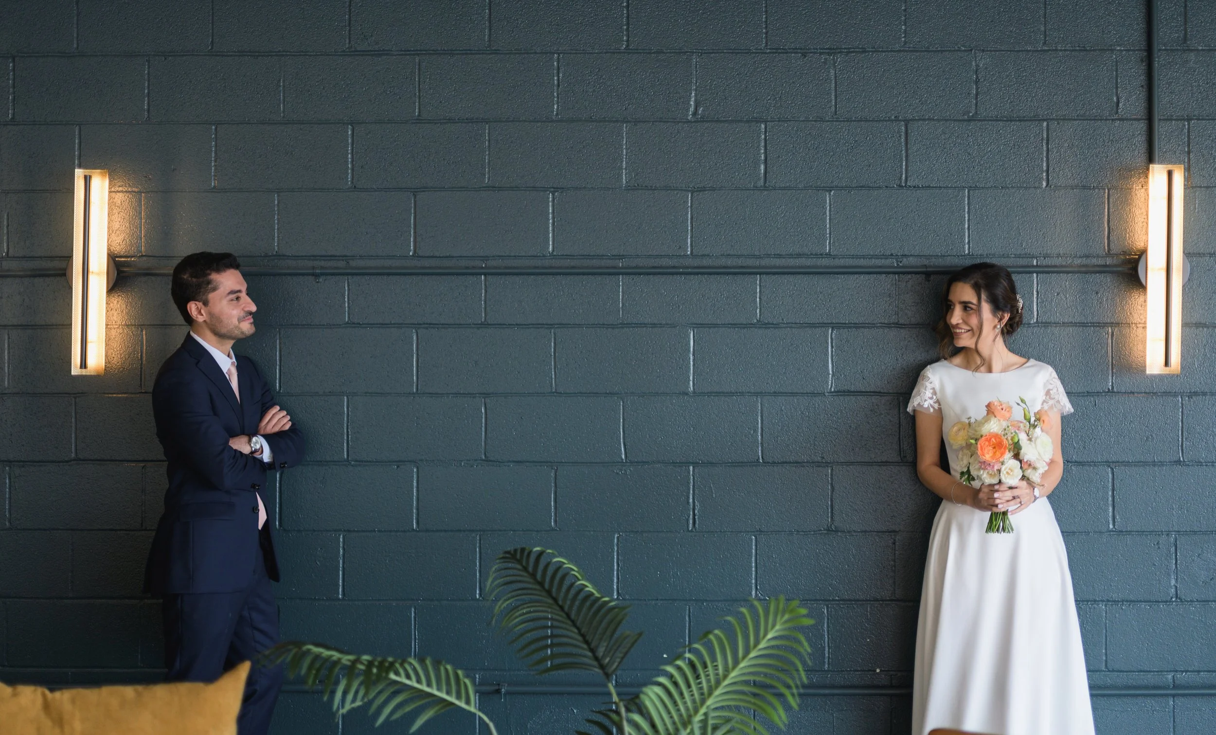 bride and groom leaning up against a blue colored wall smiling at each other. Bride is holding a bouquet, groom has his arms crossed