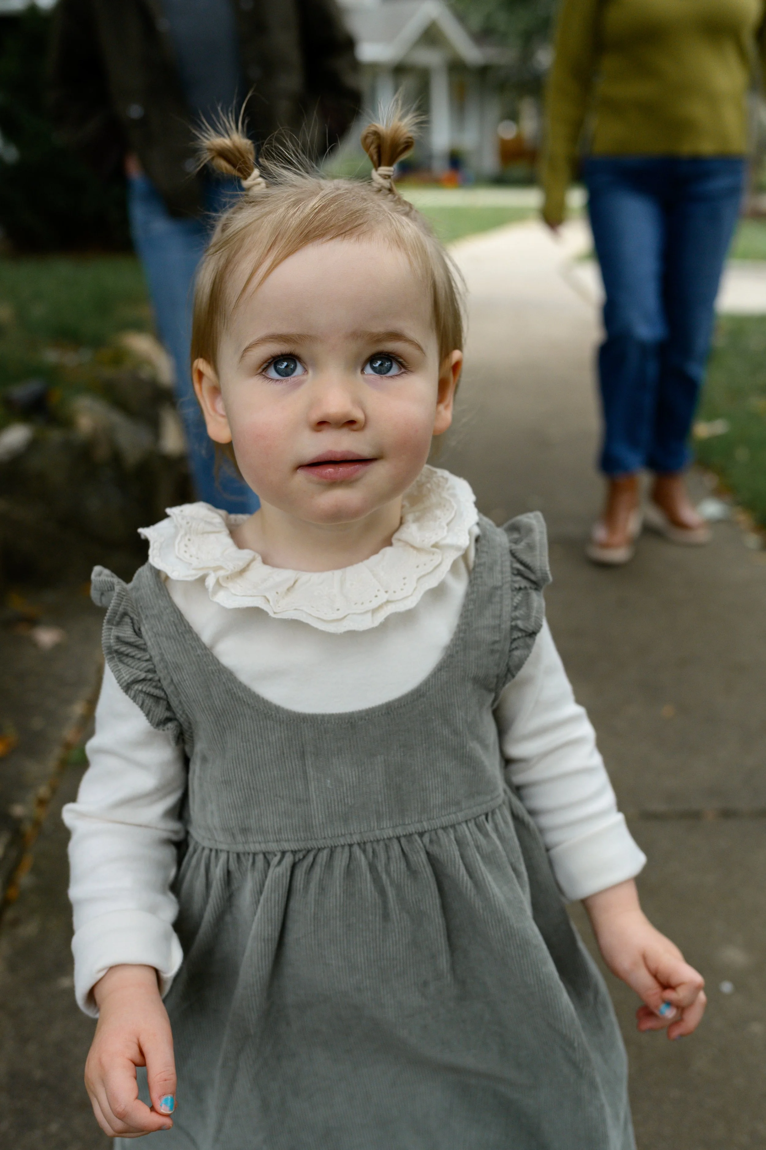 Young girl with light brown hair in pigtails, blue eyes, wearing a cream-colored ruffled collar shirt under a gray dress, standing outdoors on a sidewalk.