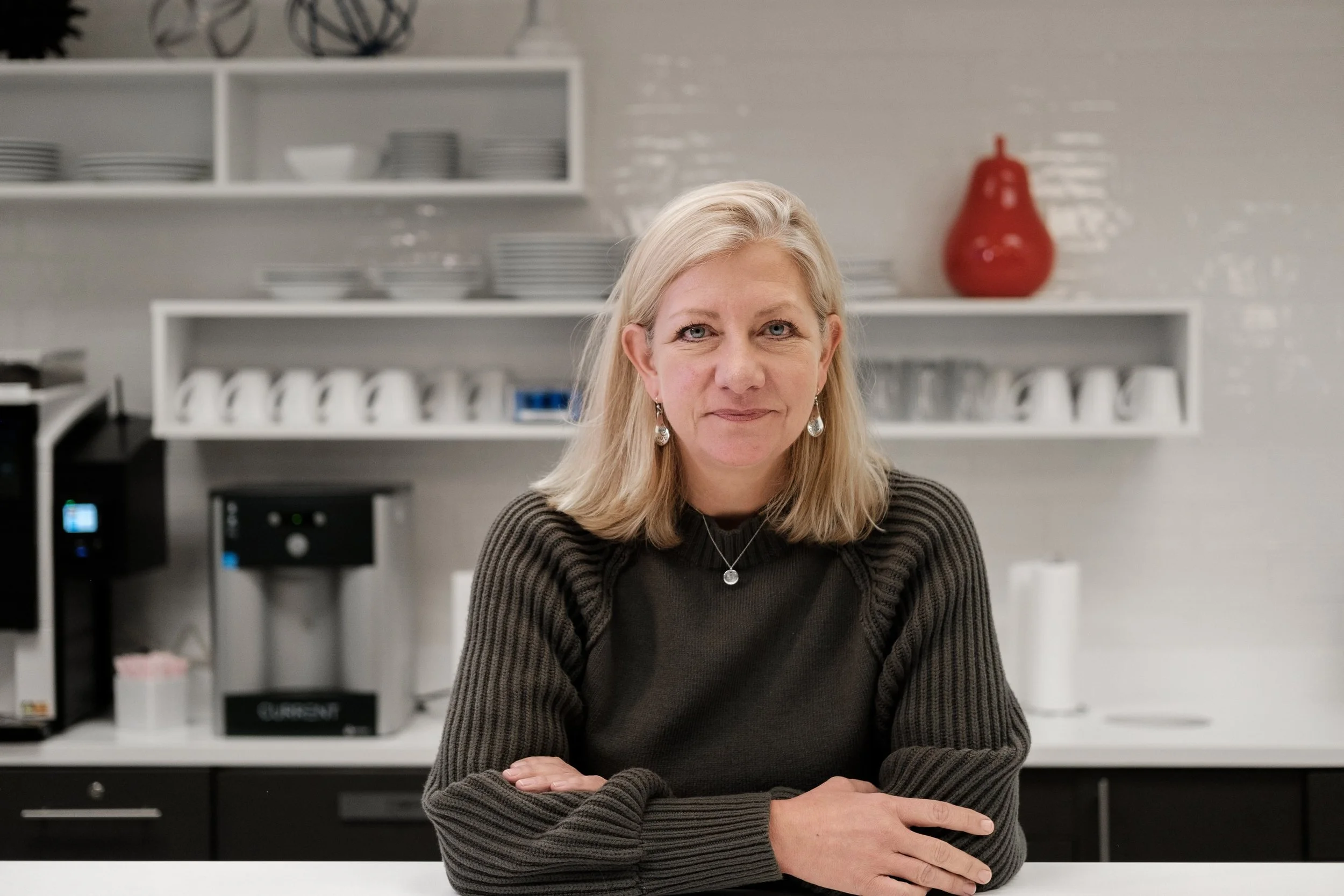 A woman with blonde hair wearing a black and gray striped sweater sitting at a kitchen counter, with a background of shelves holding plates, cups, and decorative objects, and a red vase on the wall, during a Charlotte corporate headshot photography.