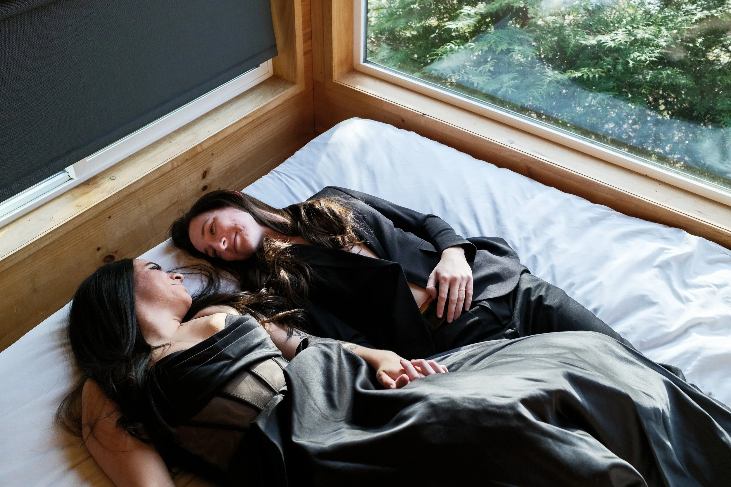 Two women lying on a bed, facing each other, in a room with large windows showing greenery outside. They are wearing dark clothing and appear to be engaged in an intimate moment.