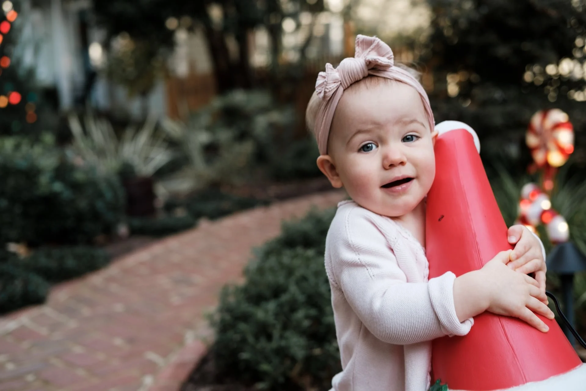 A young girl with light skin, blue eyes, wearing a light pink headband with a bow, white sweater, holding a large red Christmas-themed ornament outdoors, with Christmas decorations and greenery in the background in Charlotte, NC.