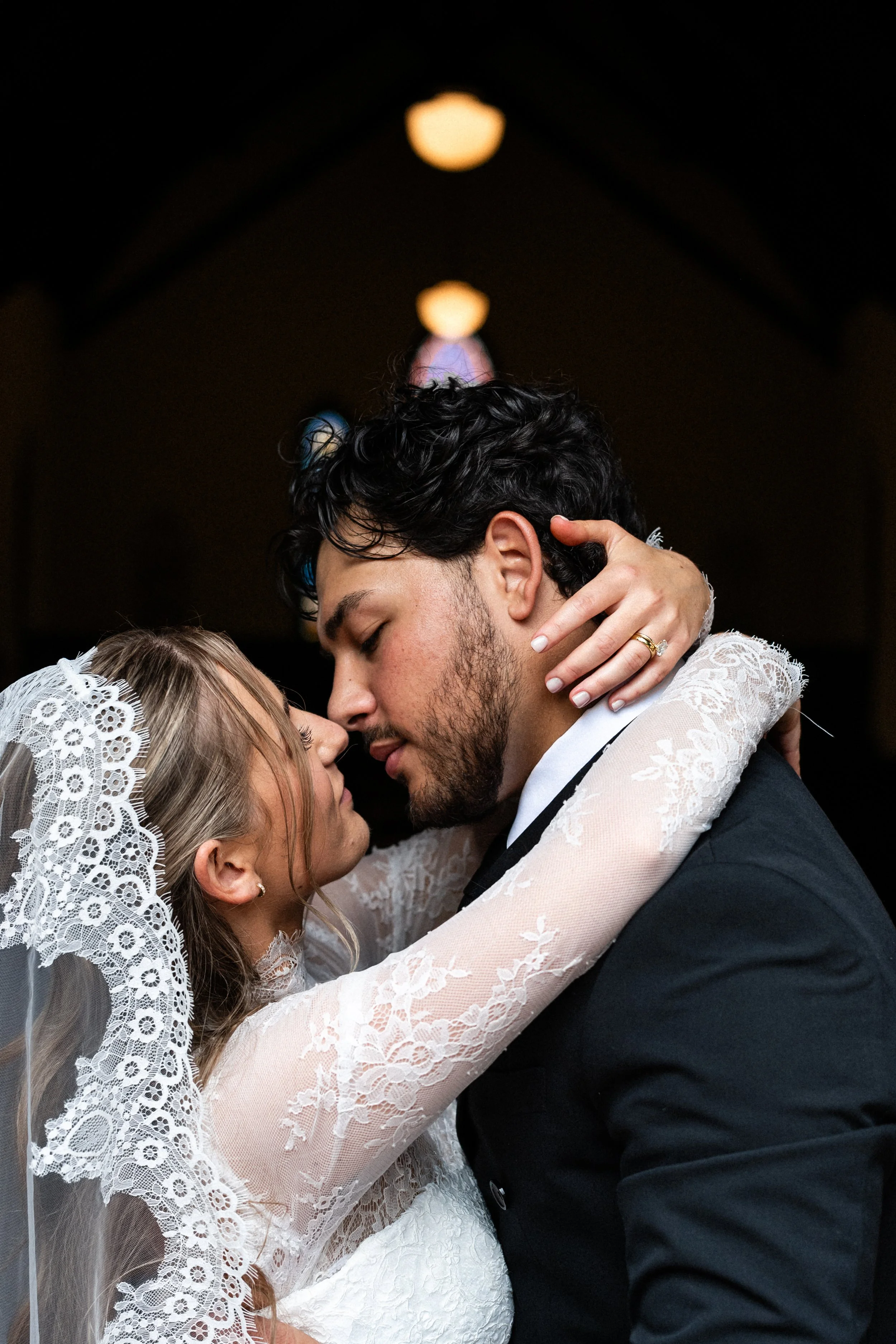 A bride and groom are close together, touching foreheads and eyes closed, in a wedding moment. The bride is wearing a lace wedding dress and veil, and the groom is in a black suit with a white shirt. The bride's hand, with a wedding ring, is gently on the groom's face