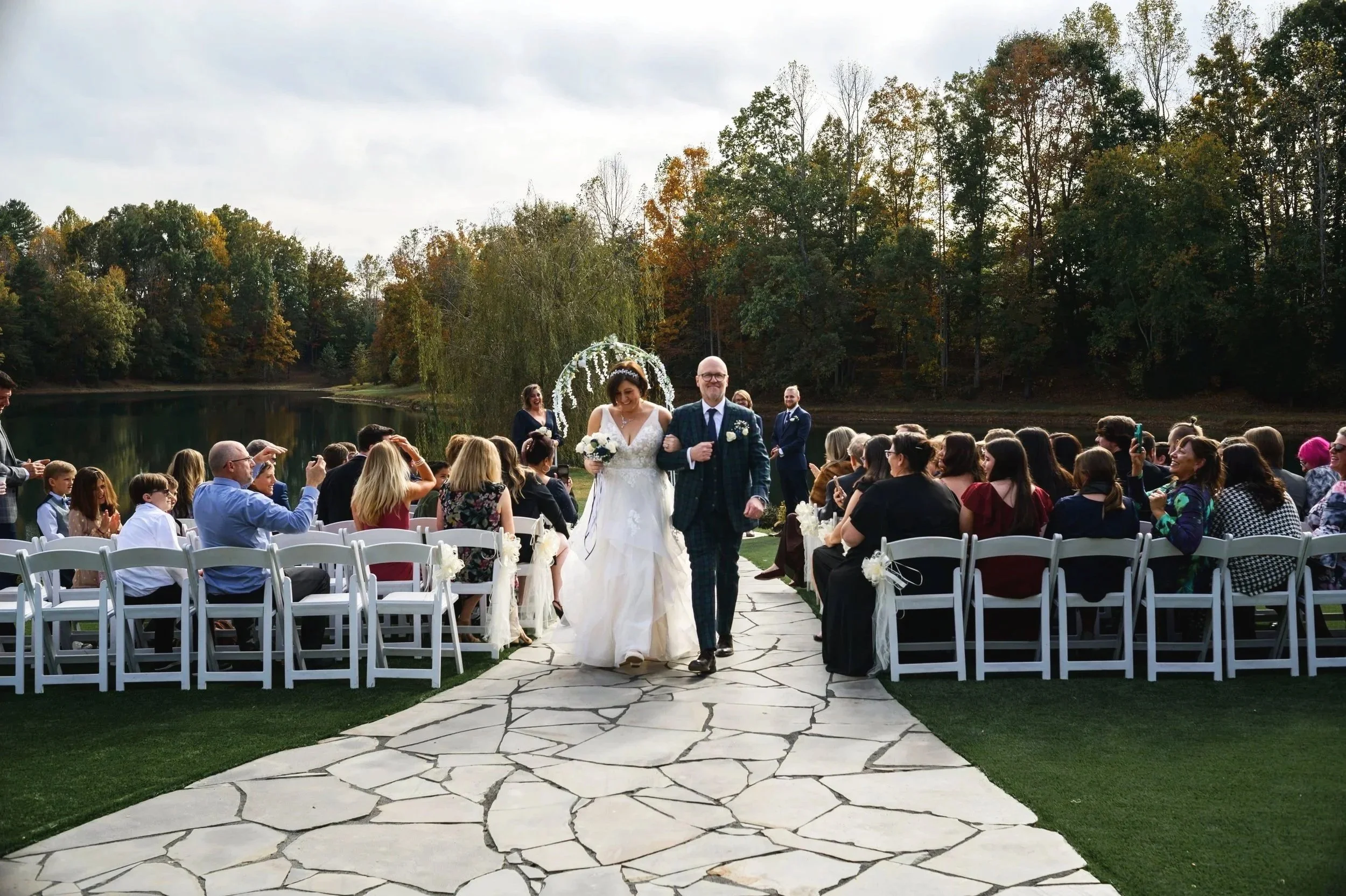 Bride and groom walking down an outdoor aisle during a wedding ceremony at Splendor Pond Flower Farm, with guests seated on either side, at a scenic lakeside setting surrounded by trees with autumn foliage.