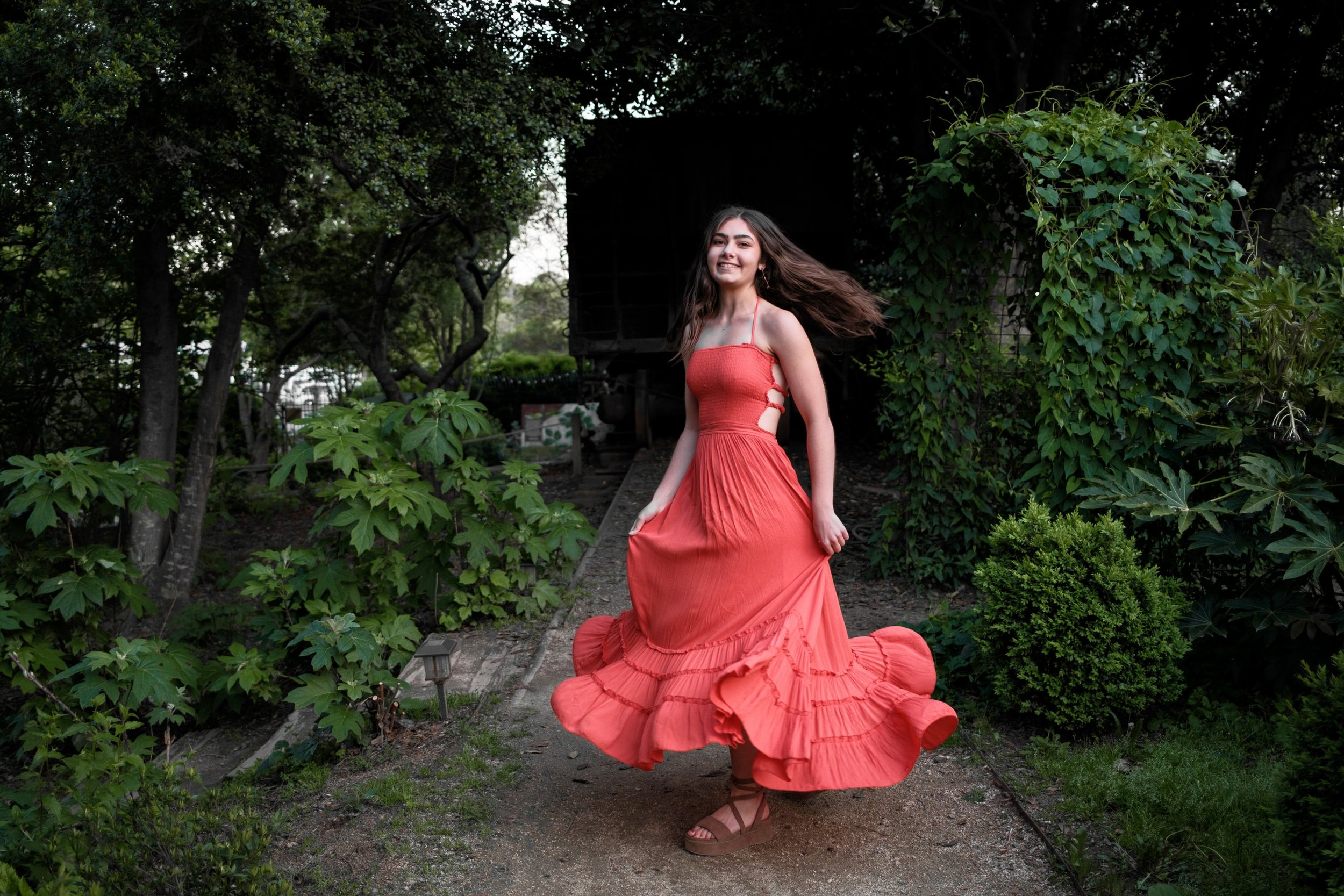 Young woman in a coral dress twirling on a garden path surrounded by lush green foliage.