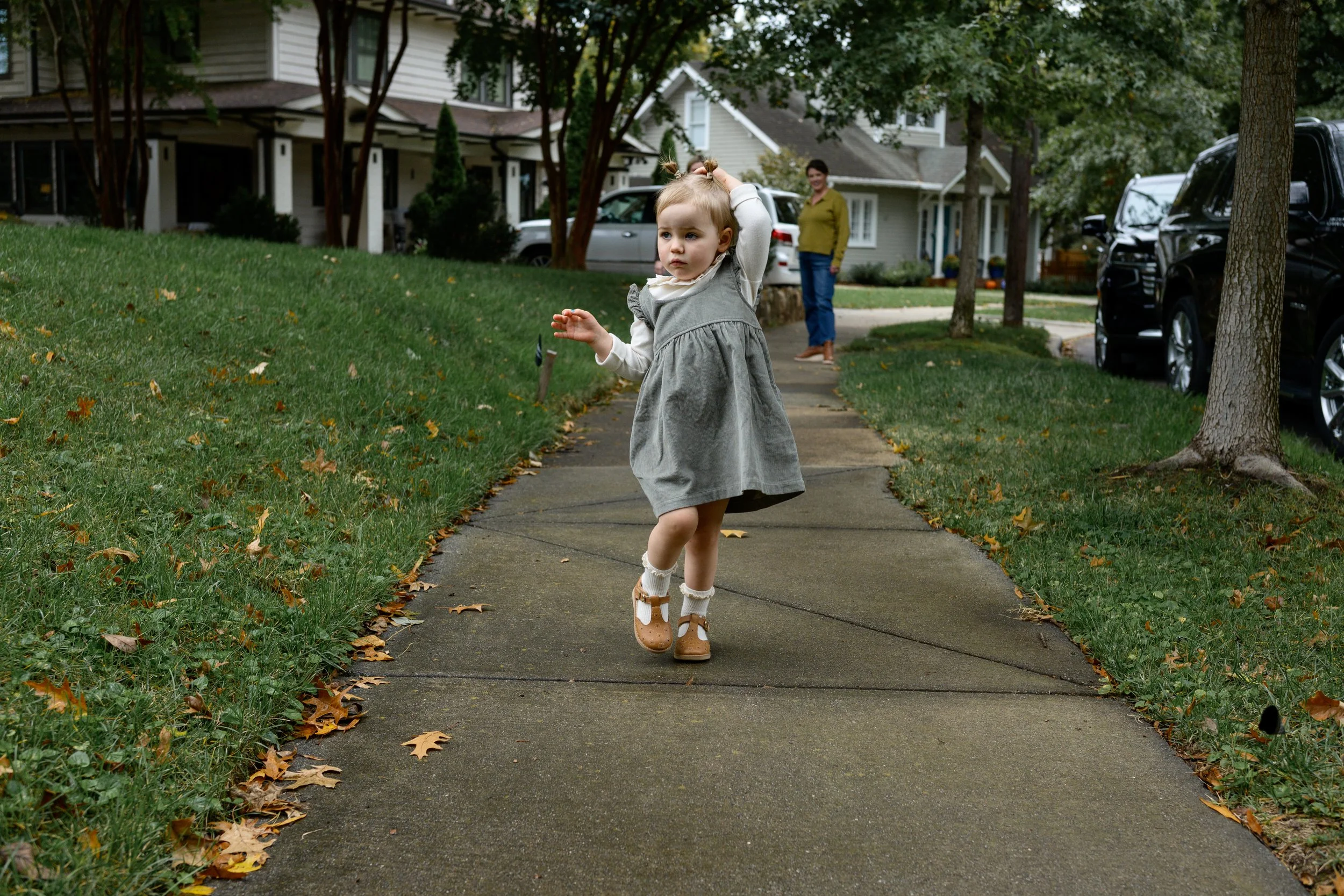 A young girl with blonde hair in pigtails wearing a grey pinafore dress, white socks, and brown shoes, running on a sidewalk while holding her hair, with an older woman watching in the background on a residential neighborhood street.