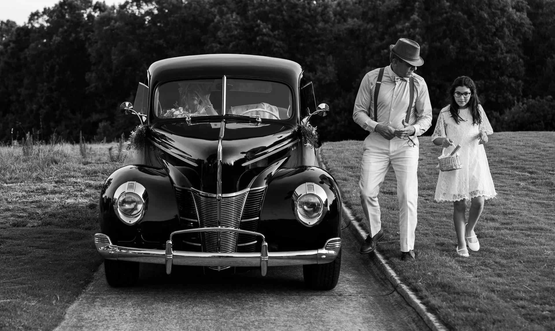 Black and white photograph of a vintage car parked on a grassy area, with a man and a girl walking beside it on a park or field, trees in the background.
