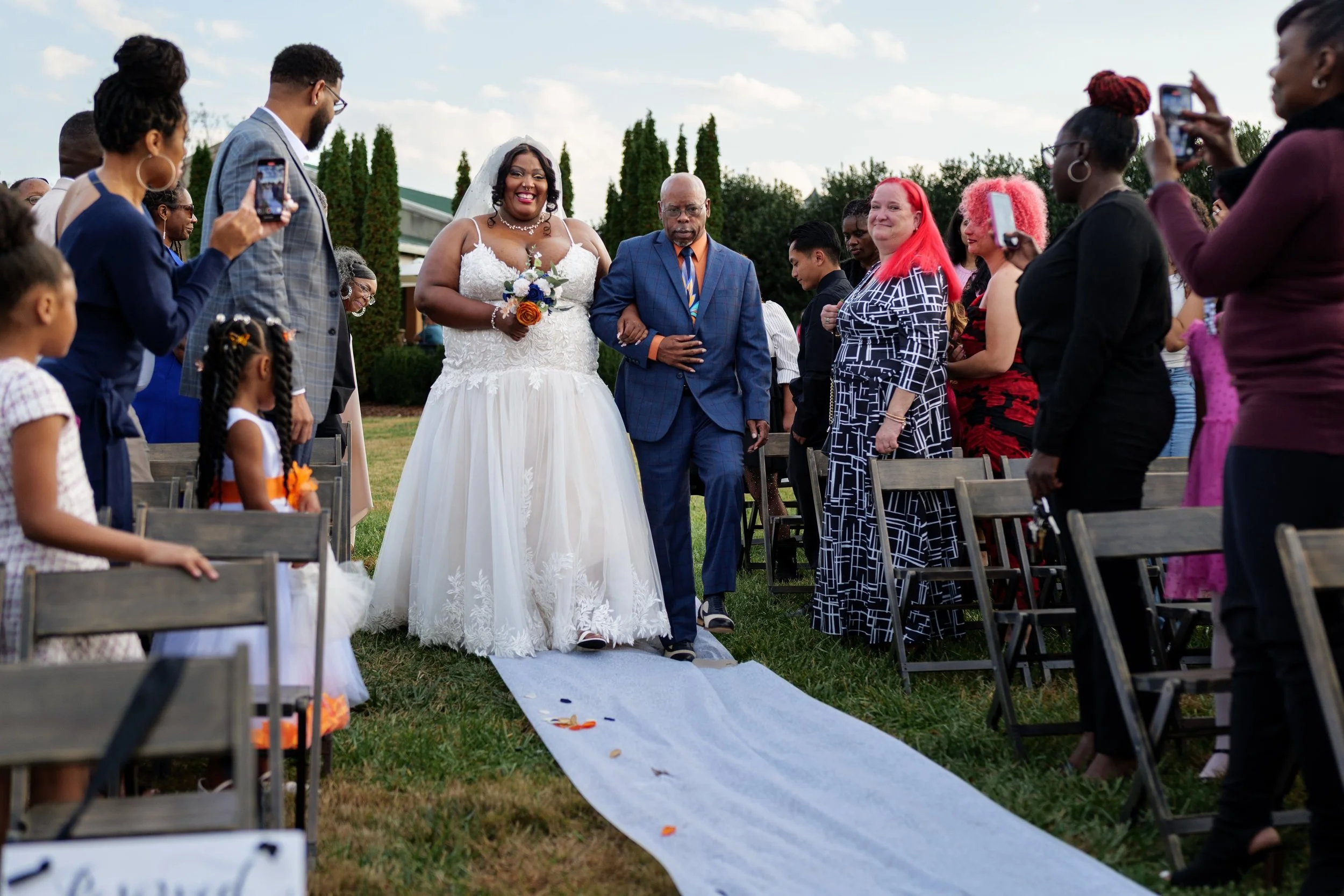 A bride walking down the aisle with her father at an outdoor wedding ceremony, surrounded by seated guests taking photos.