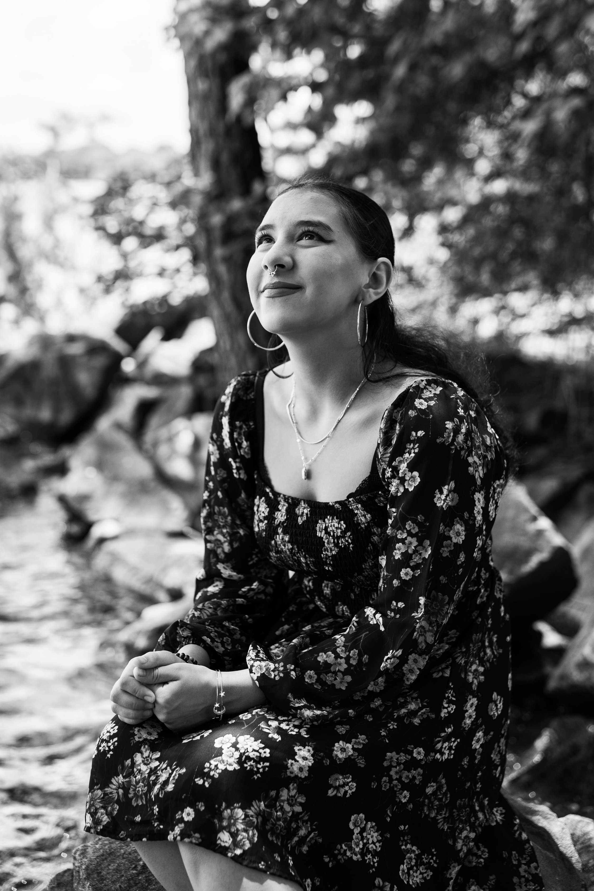 A woman with dark hair in a floral dress sitting outdoors at Jetton Park in Cornelius, NC, looking upward with a gentle smile, in black and white.