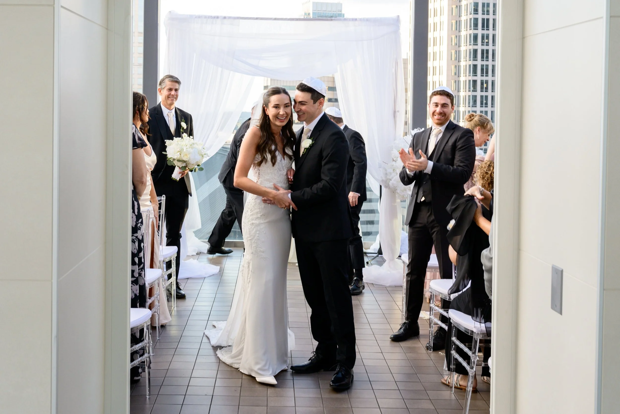 A bride and groom holding hands and smiling during their wedding ceremony at Nuvole They are dressed in formal wedding attire, with the bride in a white gown and the groom in a dark suit. Guests are seated on either side, some standing and clapping, 