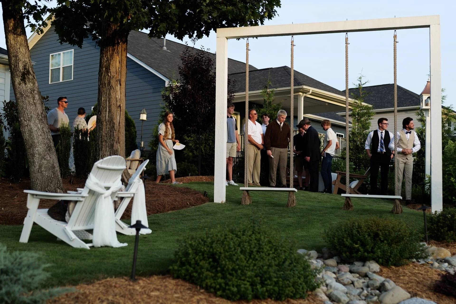 People gathered in a backyard garden with a white swing set, trees, bushes, and Adirondack chairs, possibly for a social event or gathering.