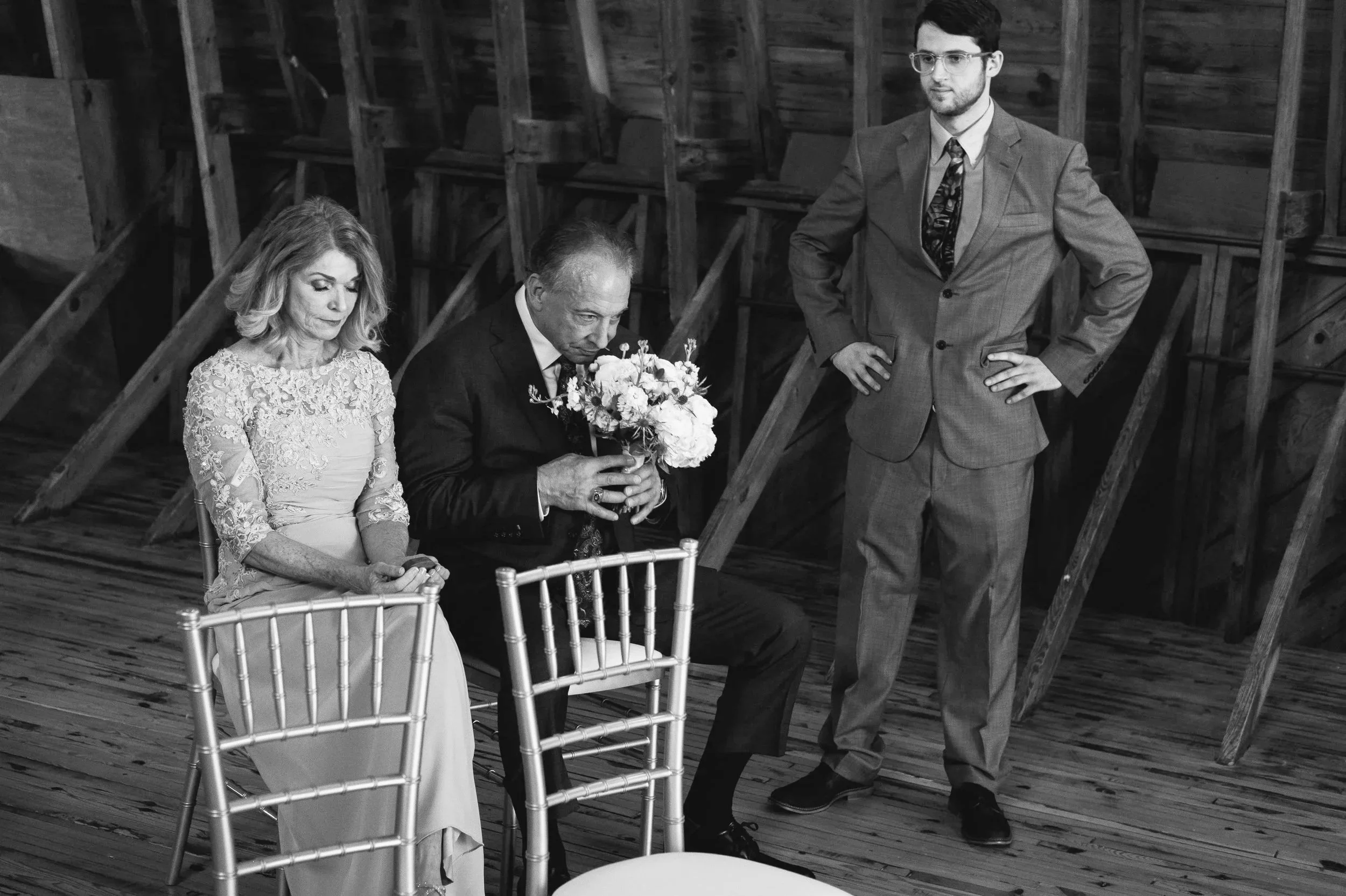 A black and white photo of a woman, an older man, and a younger man. The woman and older man are sitting with their heads bowed, possibly praying or reflecting. The woman is wearing a lace dress, and the older man is holding a bouquet of flowers. The younger man is standing with his hands on his hips, dressed in a suit, watching the scene. They are in a wooden, rustic setting, possibly a barn or similar structure.