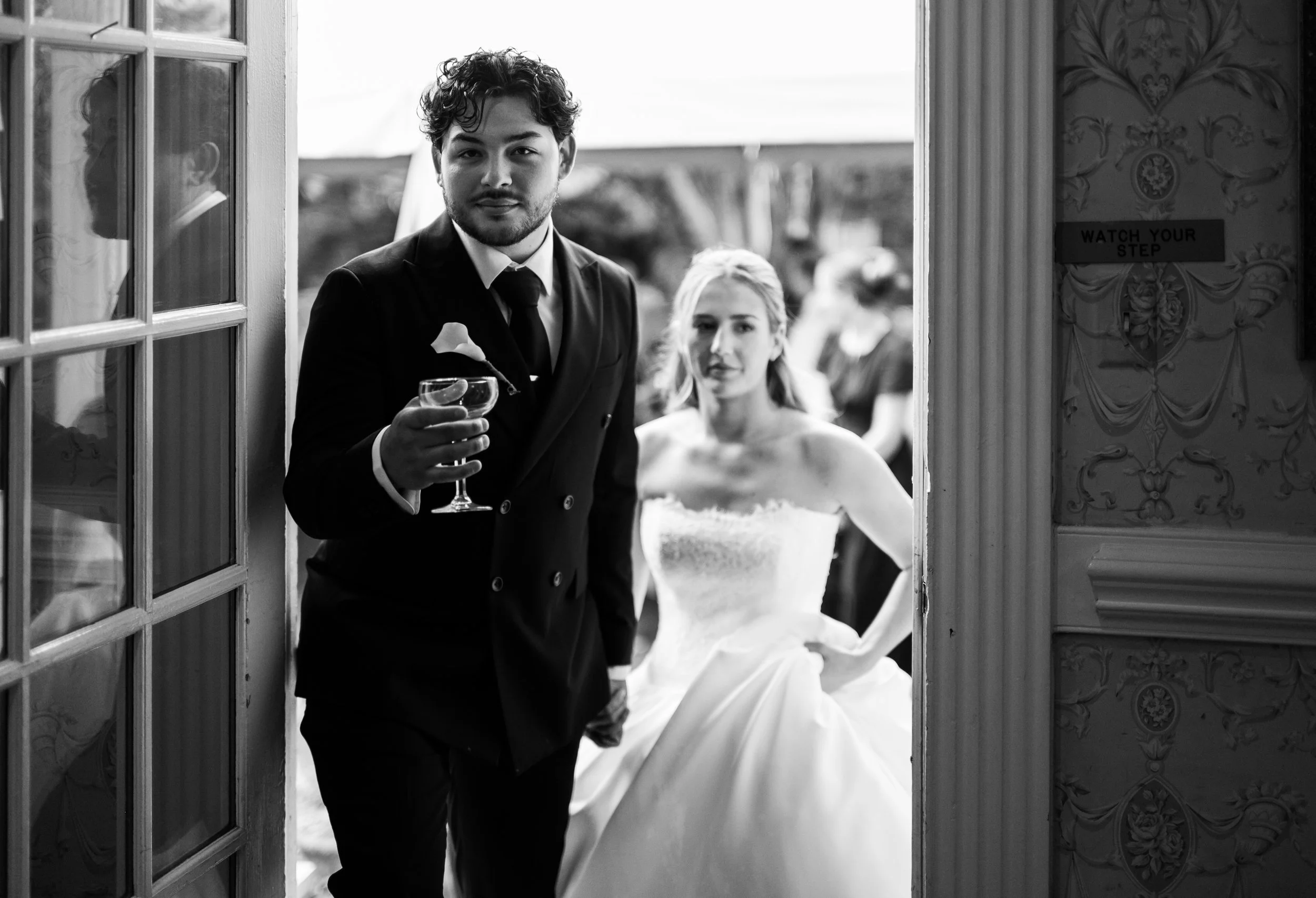 A man in a suit and a woman in a wedding dress standing in a doorway at The Morehead Inn, holding drinks, during their wedding photography.
