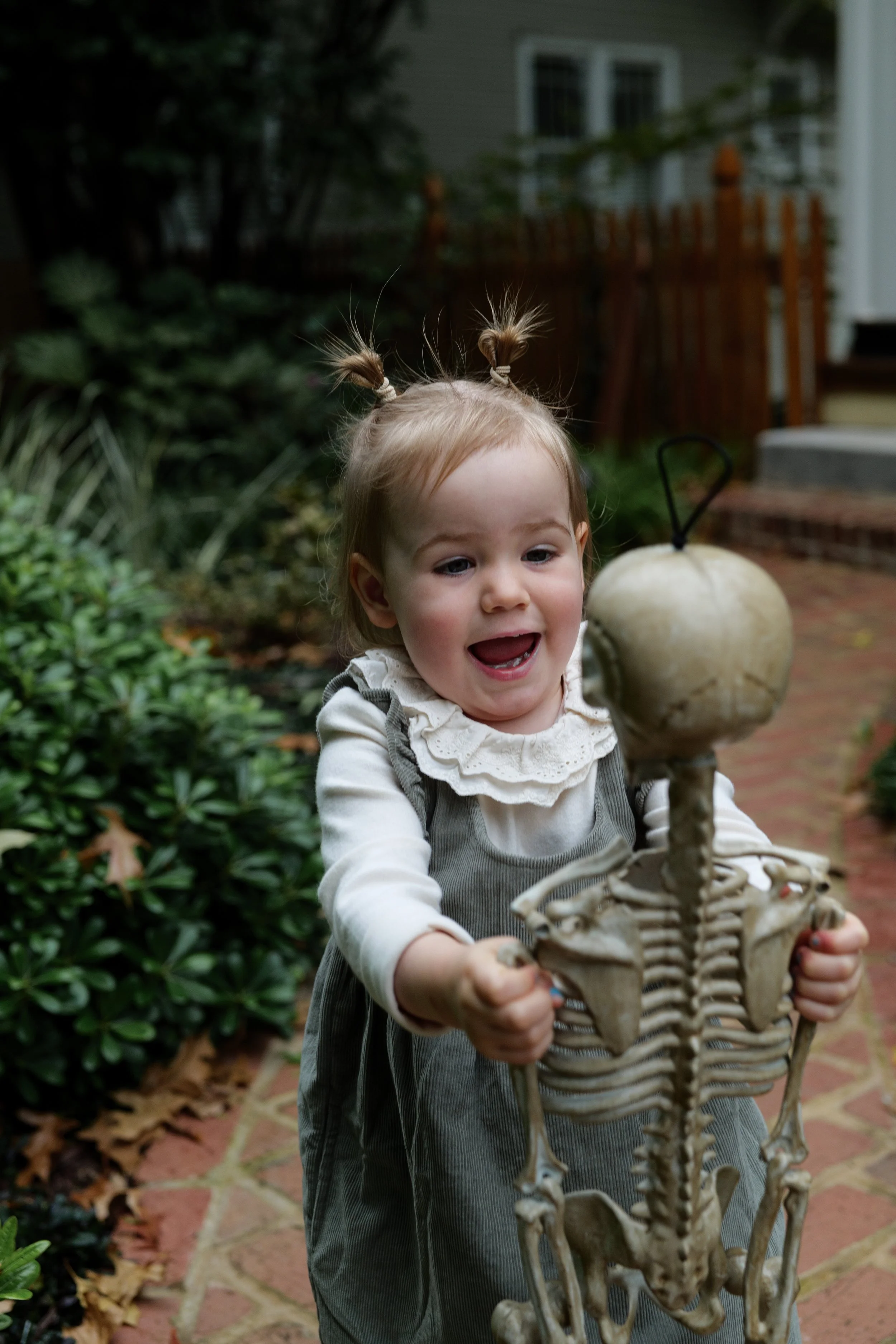A young girl with blonde hair in pigtails is smiling and holding a skeleton prop outside in a backyard.
