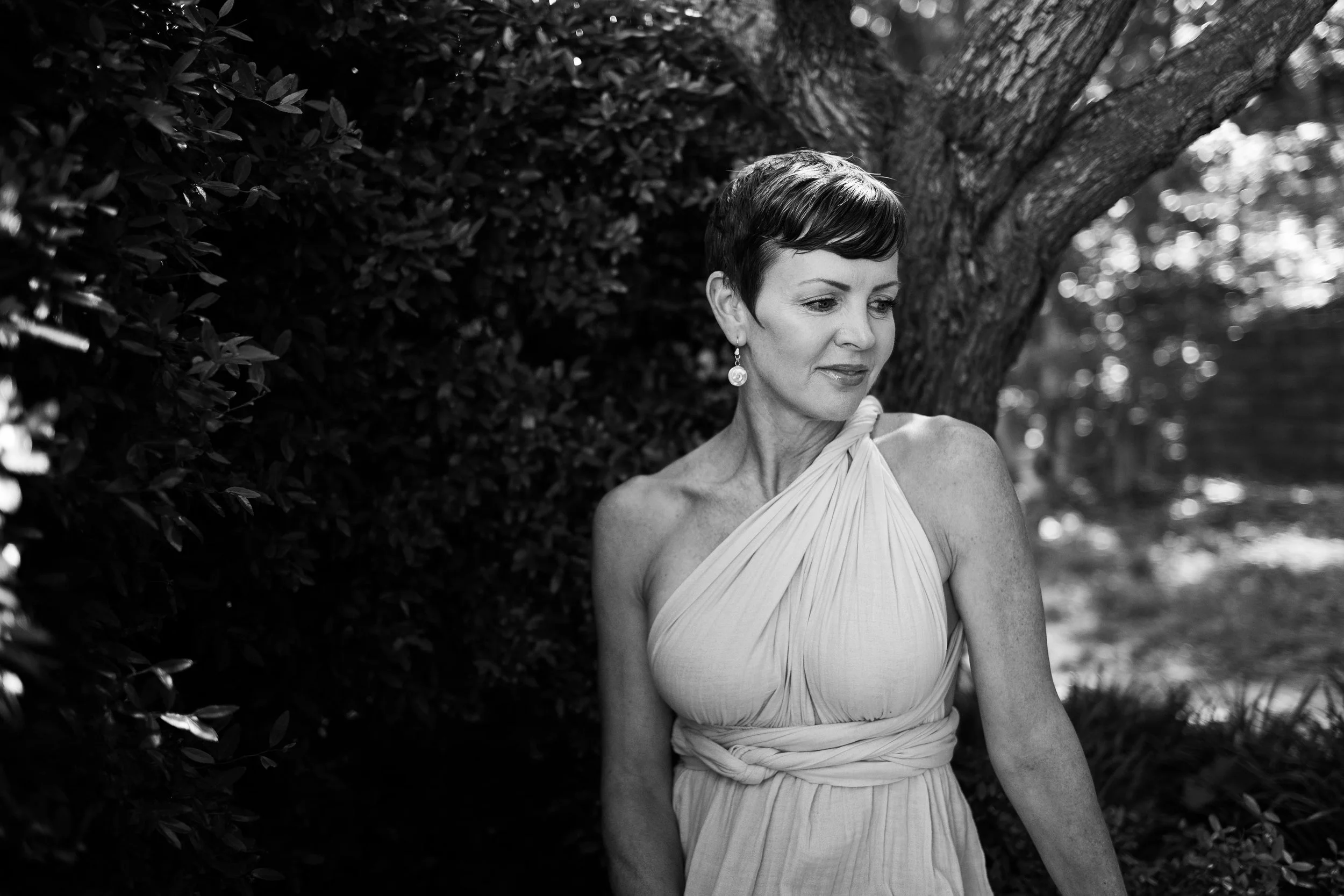 A woman with short dark hair and earrings is standing outdoors next to a large tree and bushes, looking down with a gentle expression.