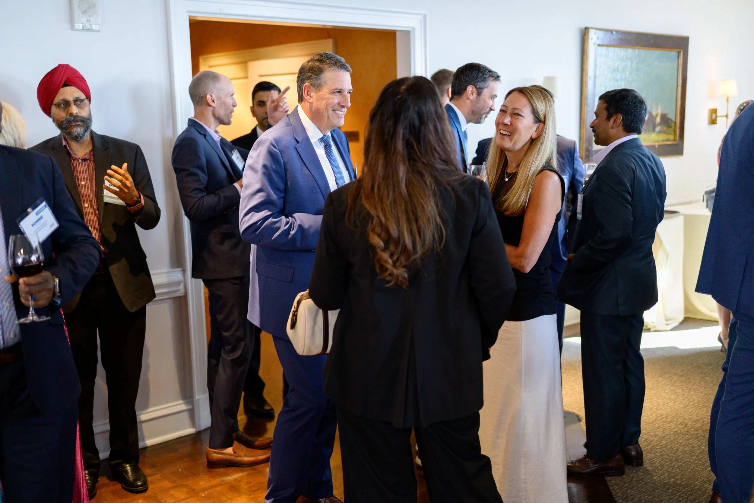 A group of professionals at a networking event, talking and laughing, in a well-lit indoor setting with artwork on the wall.