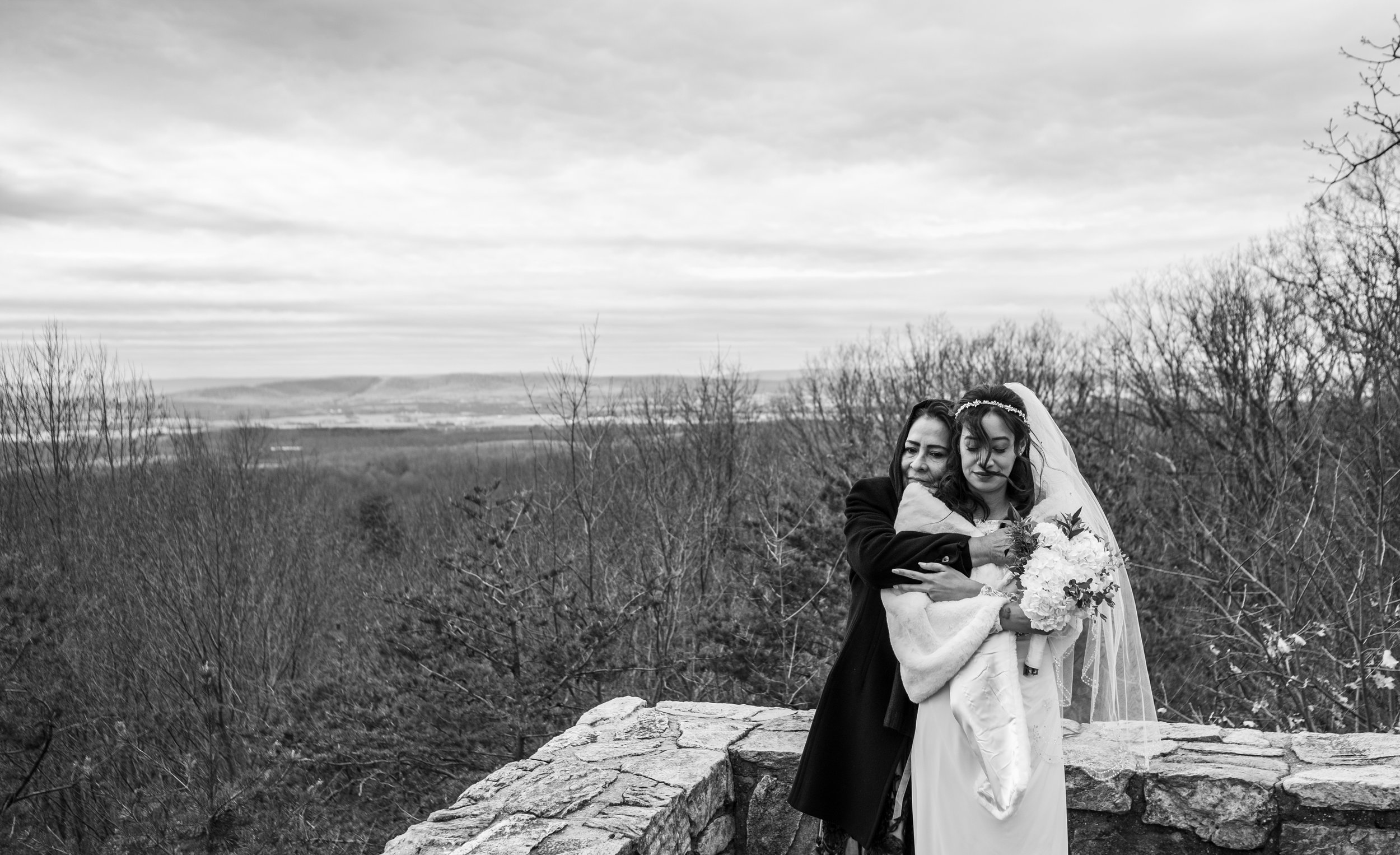 A black and white photo of two women, one dressed as a bride with a wedding dress and veil, and the other in dark clothing, hugging outdoors near a stone wall with sparse trees, an overcast sky, and distant hills.