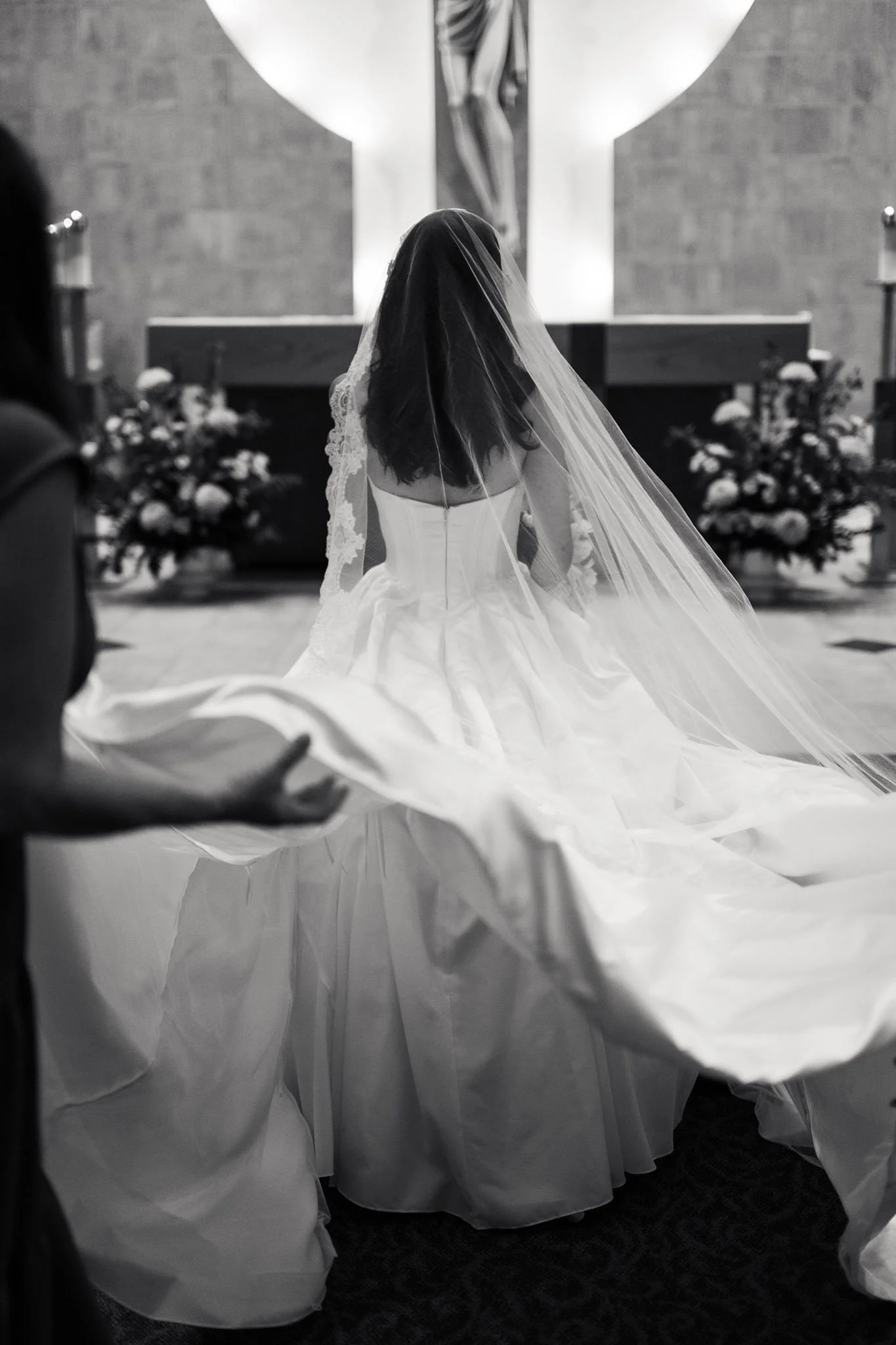 Black and white photo taken inside St. Mathews Catholic church during a wedding ceremony. The bride, with long dark hair and a veil, . She wears a strapless white wedding dress. The altar in the background has a crucifix and floral arrangements.