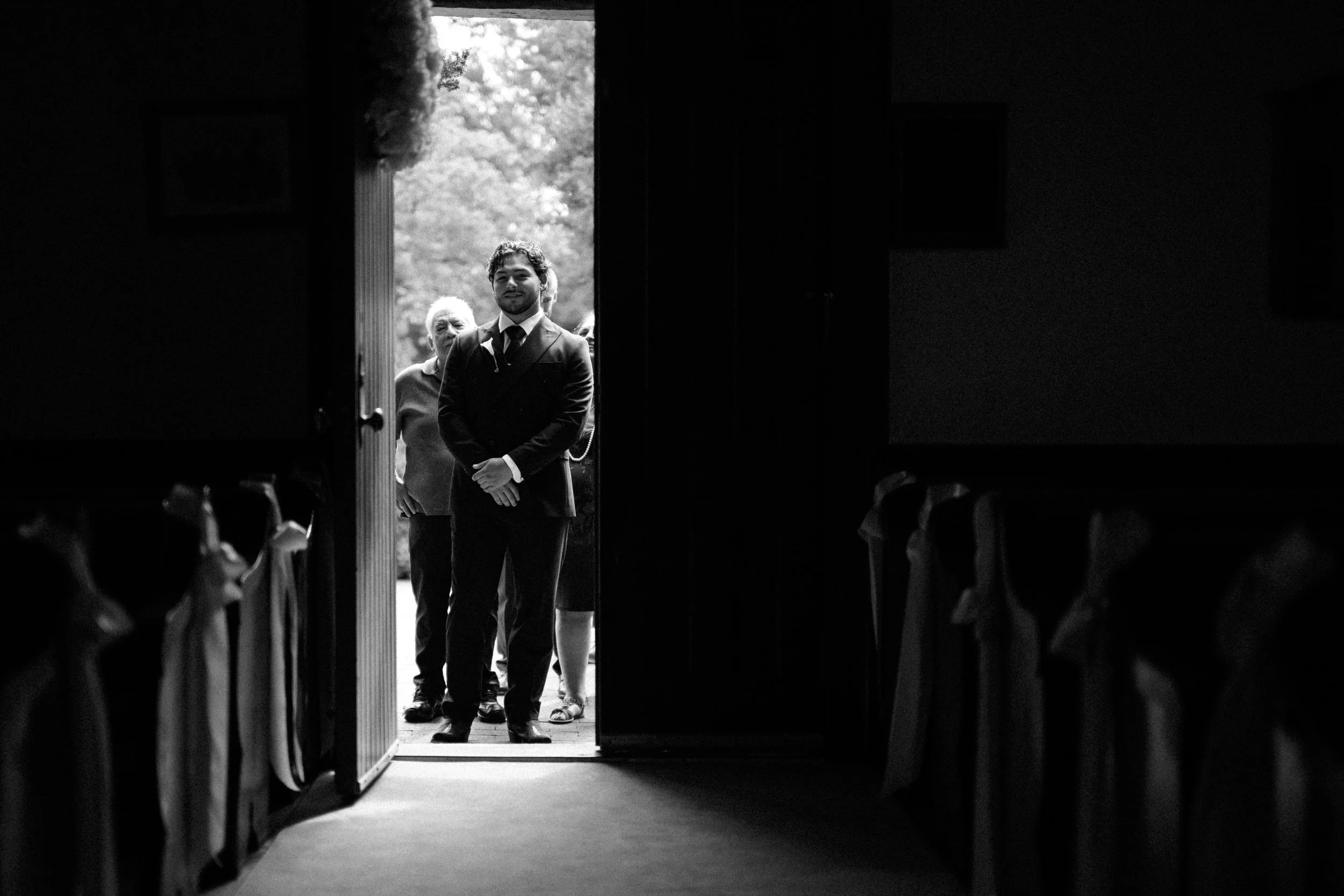 A groom in a suit and tie standing outside a chapel, with an older woman behind him, seen through a doorway from inside St. Mary's Chapel in Charlotte
