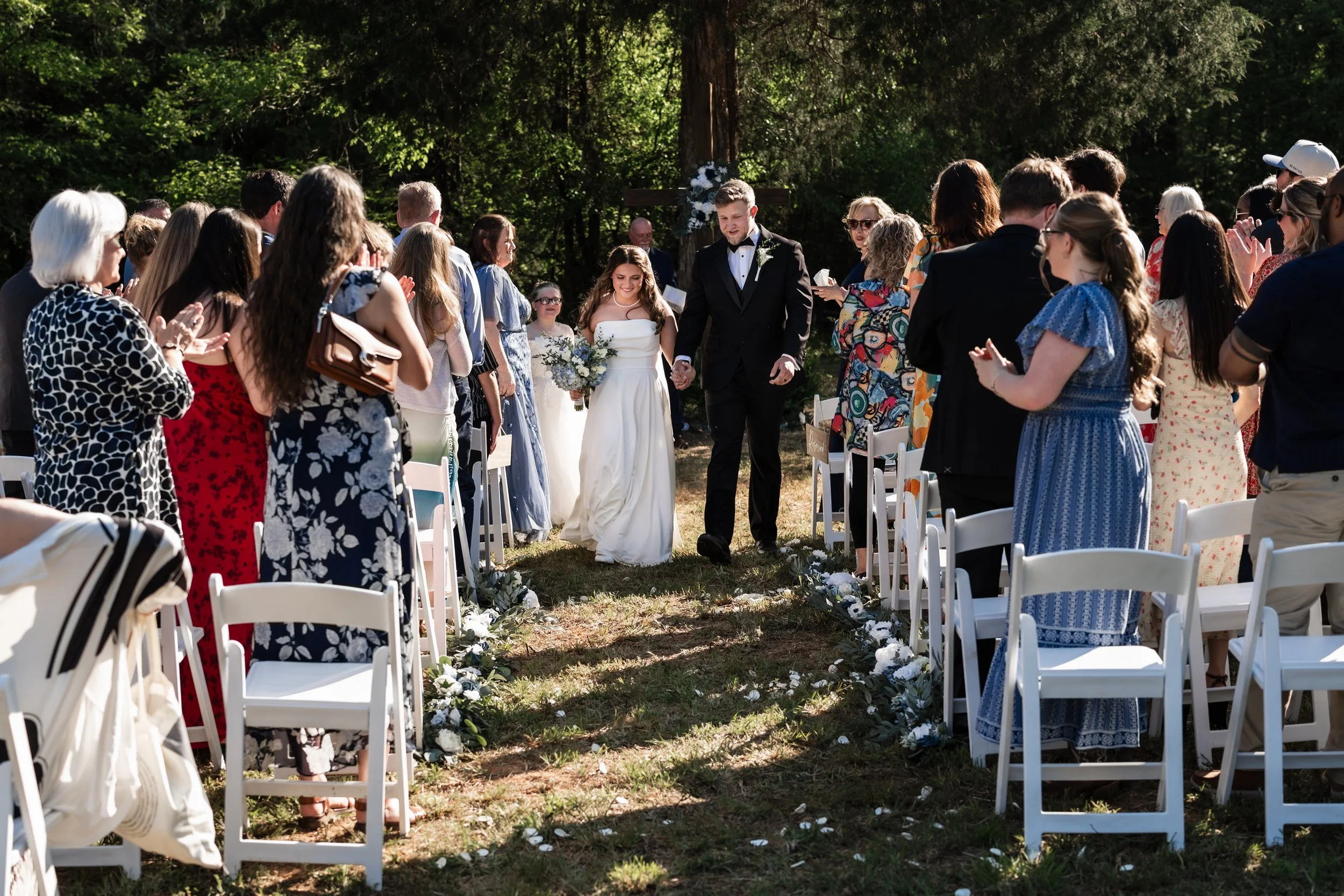 A couple getting married outdoors surrounded by guests, holding hands and walking down the aisle with floral decorations, in a wooded area with sunlight filtering through trees.