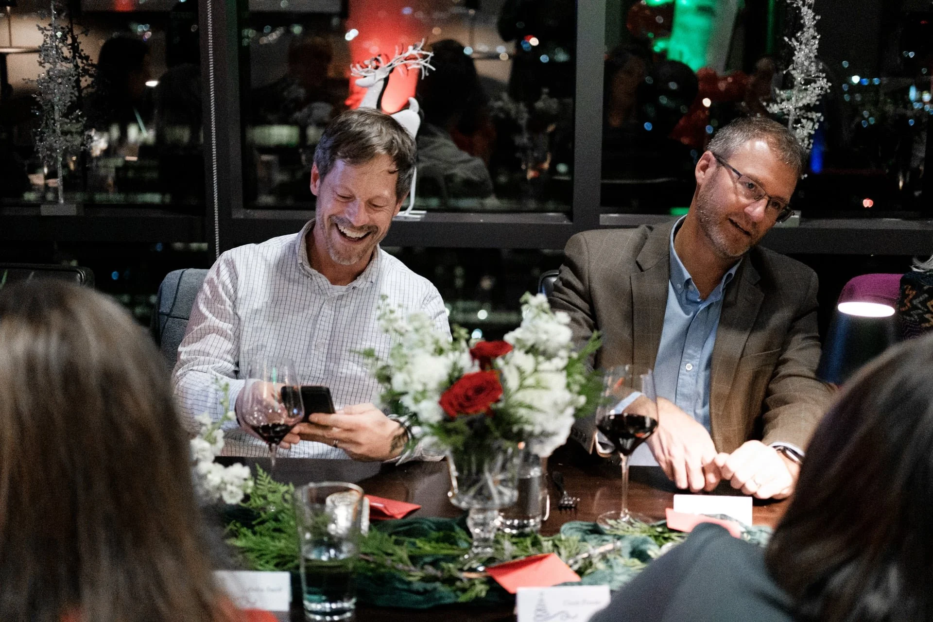 two man sitting at a table smiling during their holiday work party. One man is smiling and looking at his phone