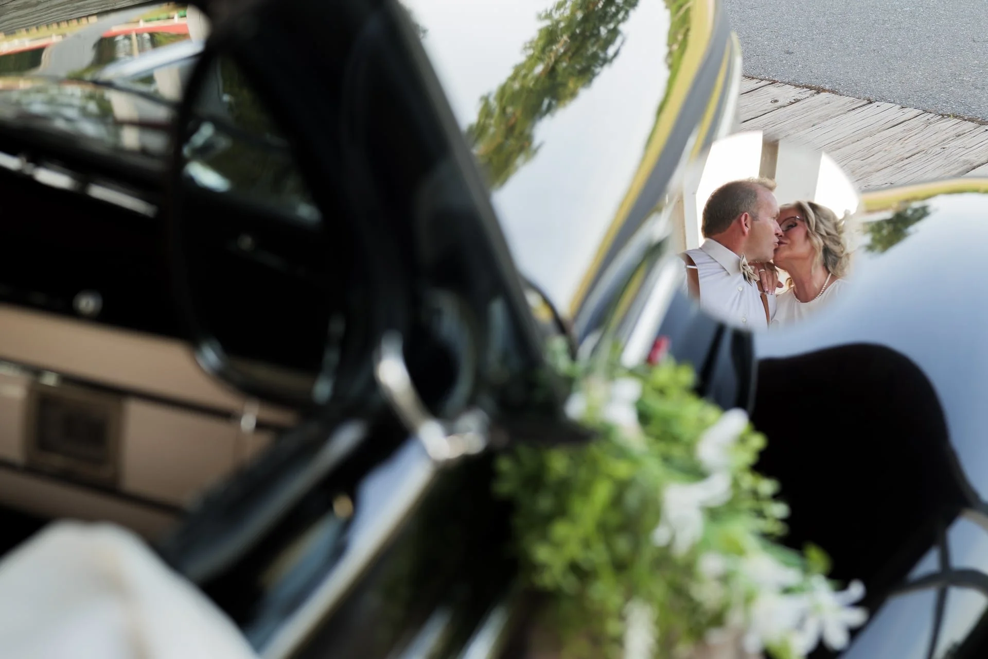 A couple kissing, reflection seen in the side mirror of a classic black car decorated with white flowers.