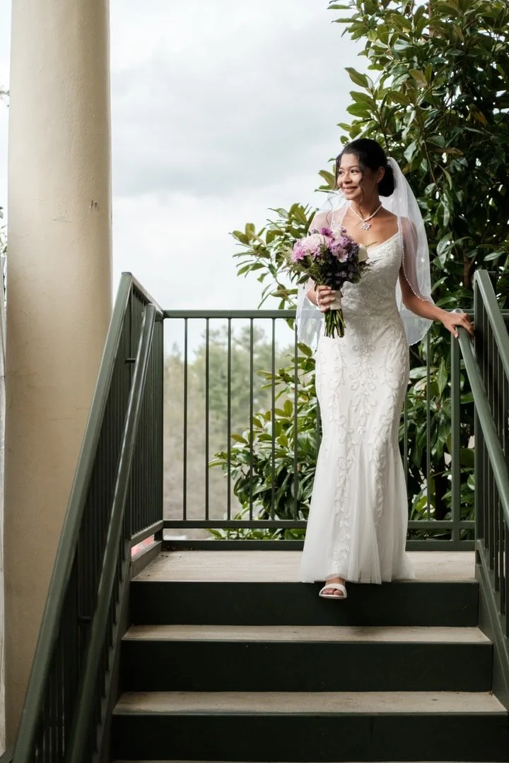 A bride in a white wedding gown holding a bouquet of pink and purple flowers, standing on a set of outdoor steps, smiling, with green trees and cloudy sky in the background.