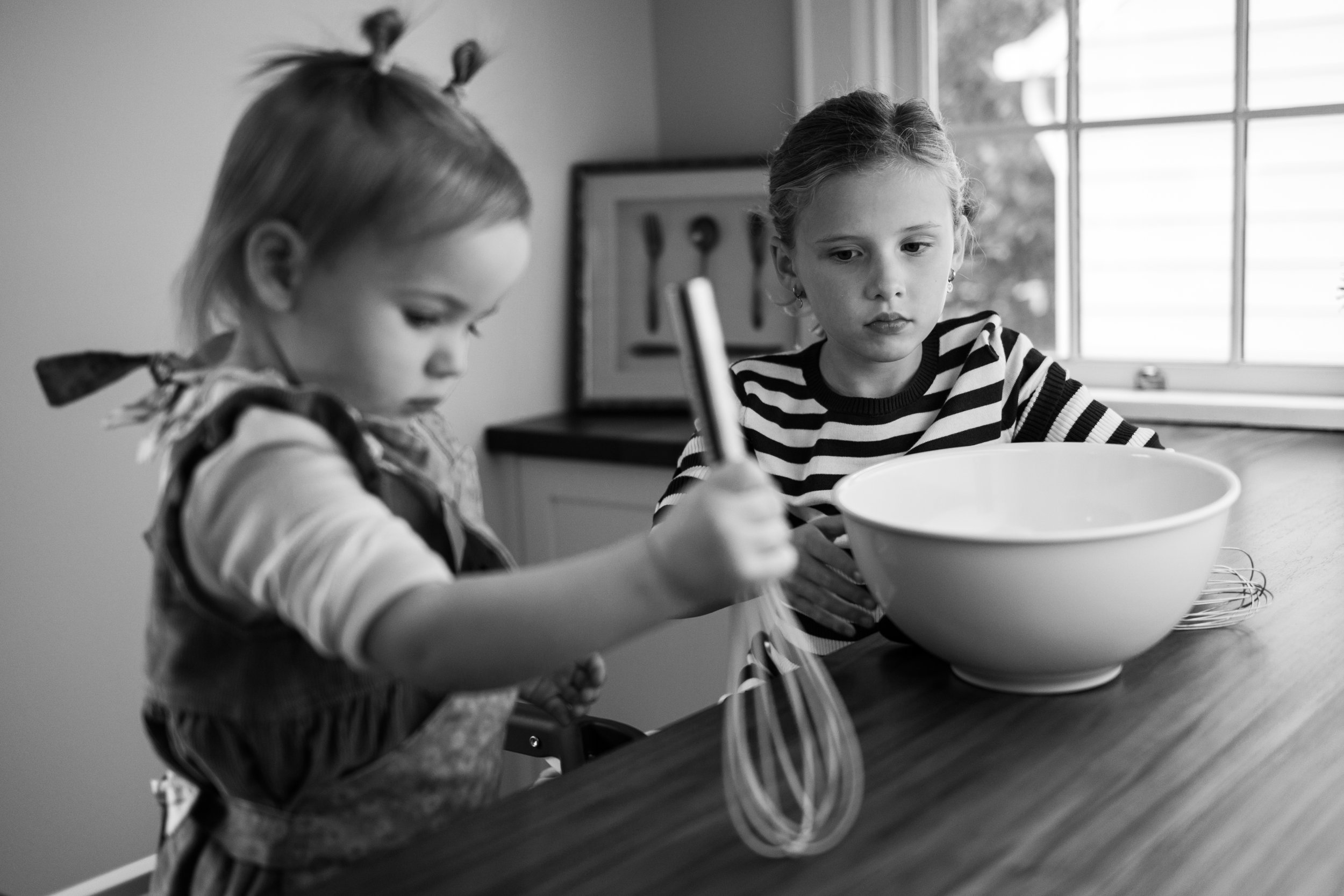Two young girls baking together at a kitchen counter, with one stirring a bowl and the other holding a whisk.