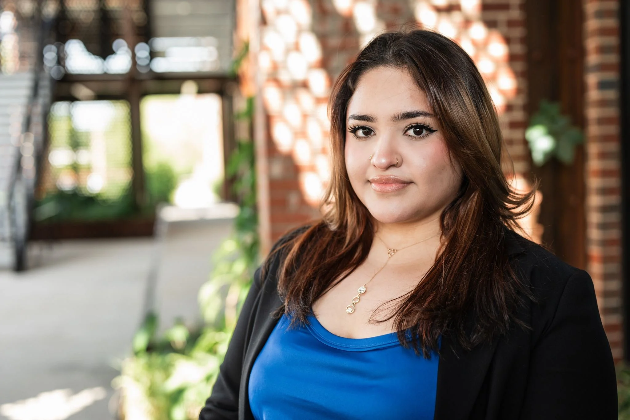 A young woman with long brown hair, silver jewelry, black eyeliner, and light makeup, standing outdoors in front of a blurred brick wall and greenery, wearing a blue top and a black jacket.