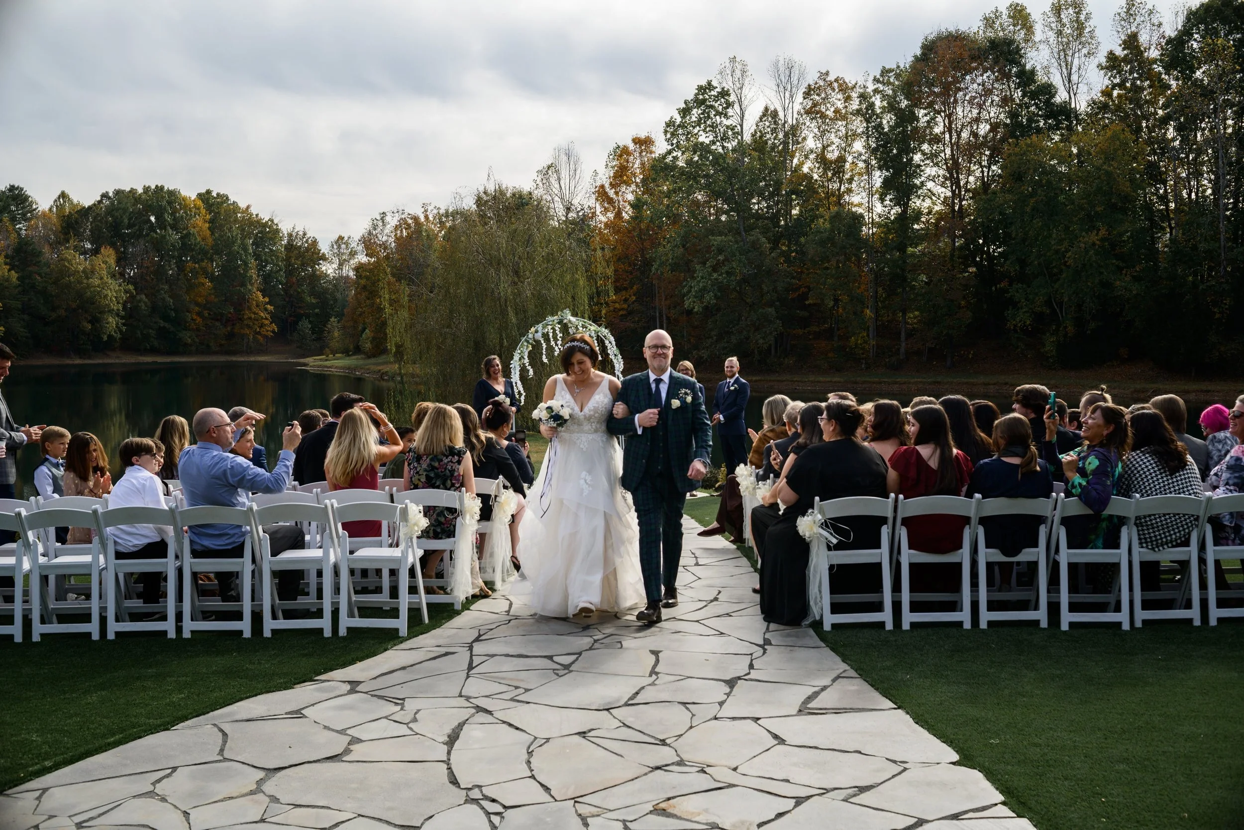 Bride walking down the aisle with her father at an outdoor wedding ceremony by a lake, guests seated on either side, trees in the background, overcast sky.