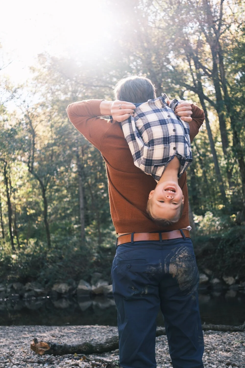 A man holding a smiling upside-down boy in a plaid shirt in a forest with sunlight filtering through the trees.