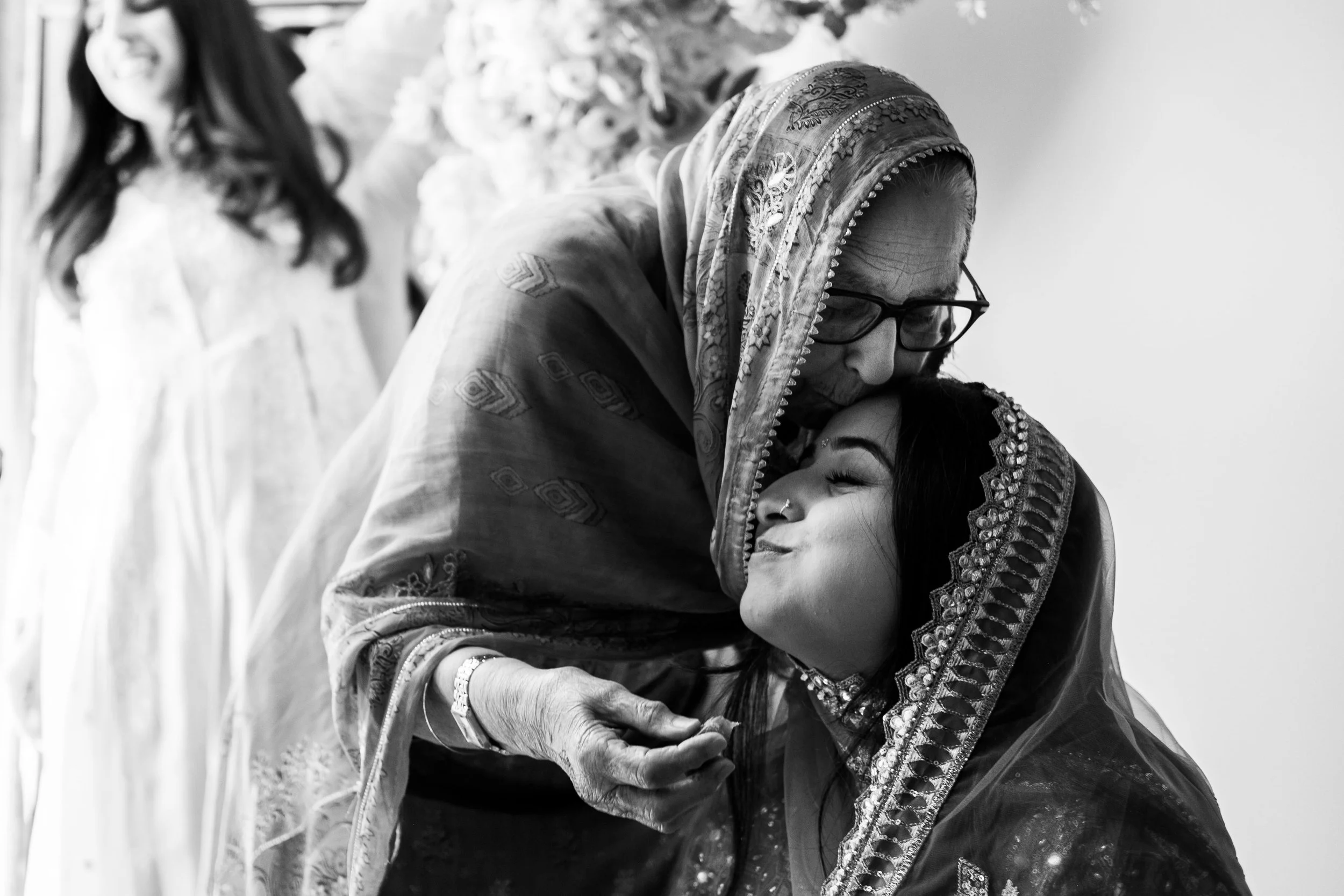 An elderly woman, wearing glasses and a traditional embroidered headscarf, shares an affectionate hug with a young woman dressed in traditional attire, smiling with eyes closed at a Charlotte Haldi and Choora ceremony