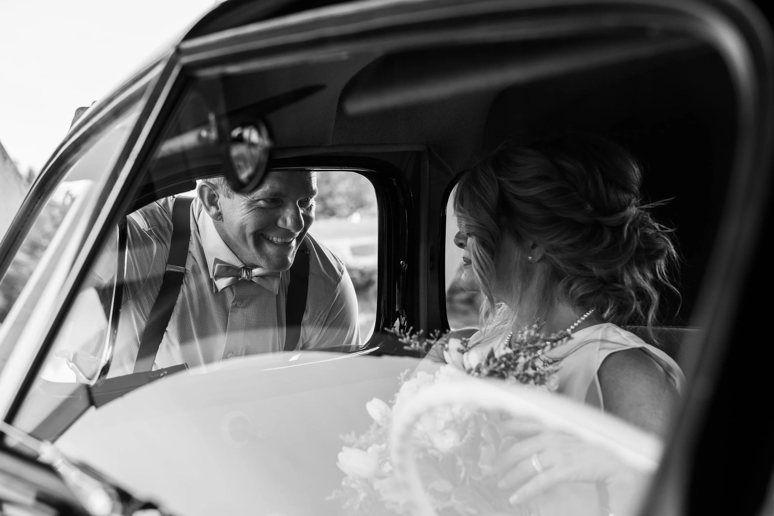 A black-and-white photo of a bride and groom in a vintage car, with the groom leaning in to kiss the bride, who is holding a bouquet, during a wedding celebration.