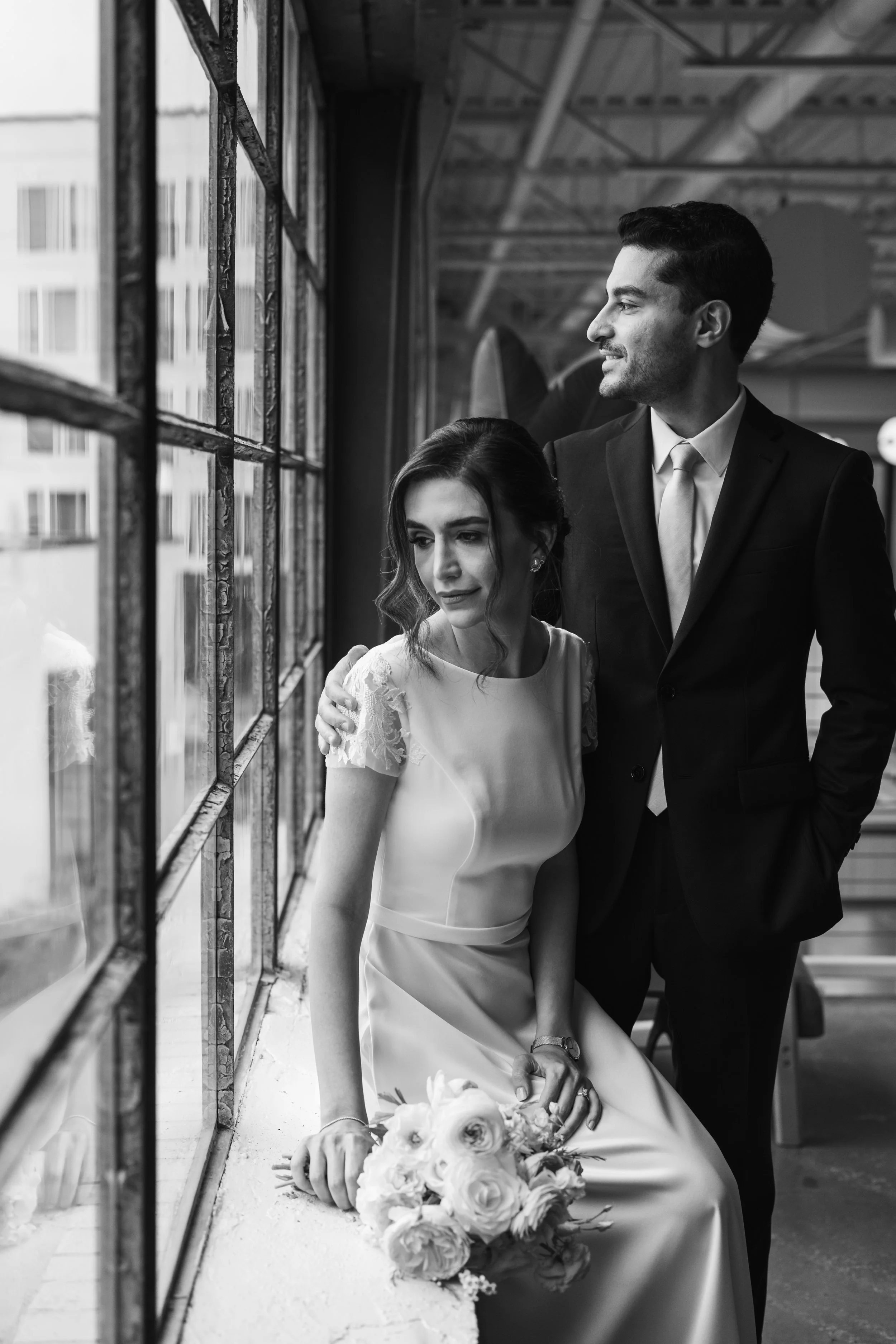 Black and white photo of a bride in a white dress sitting on a ledge and looking out a big industrial window. Her husband stands behind her at Pine venue in NoDa, Charlotte
