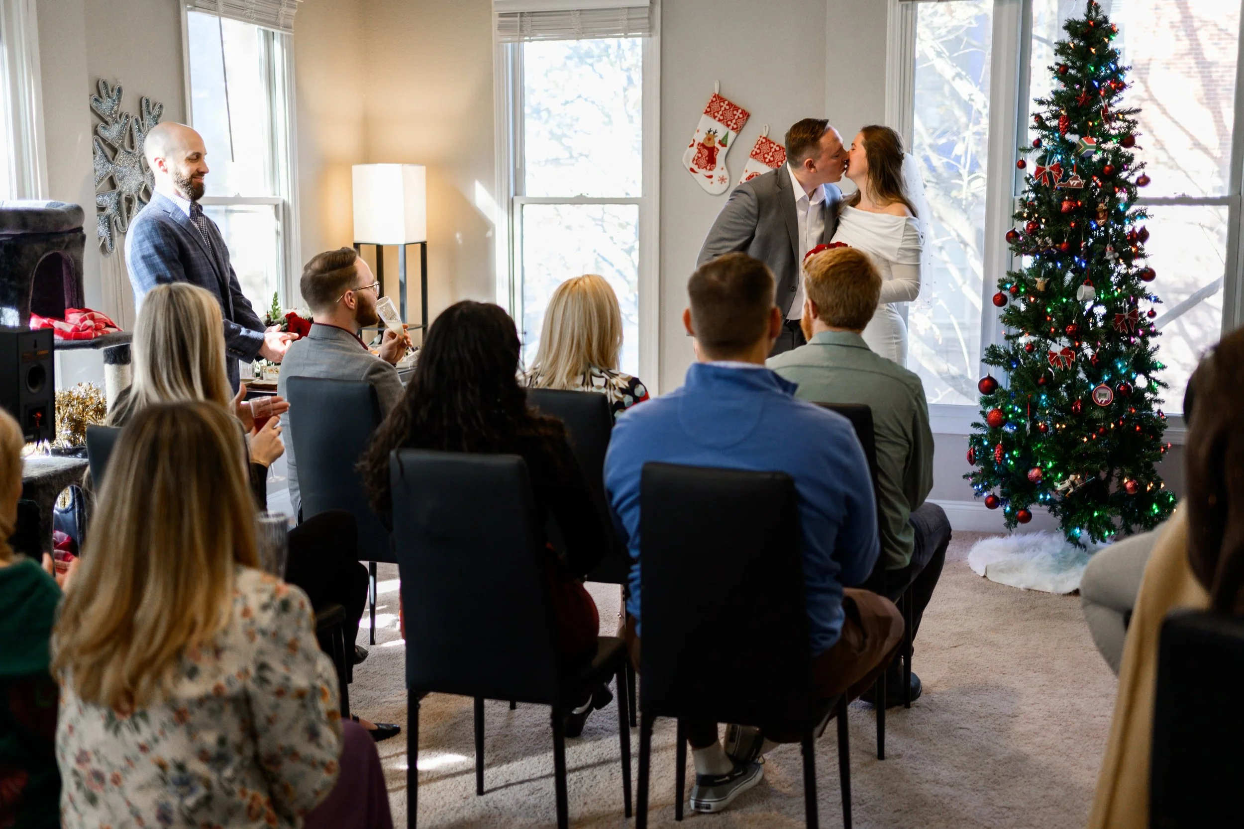 A group of people gathered in a living room decorated for Christmas, with two people kissing near a Christmas tree, while others watch and smile. Some are seated, holding drinks, and one person stands nearby.
