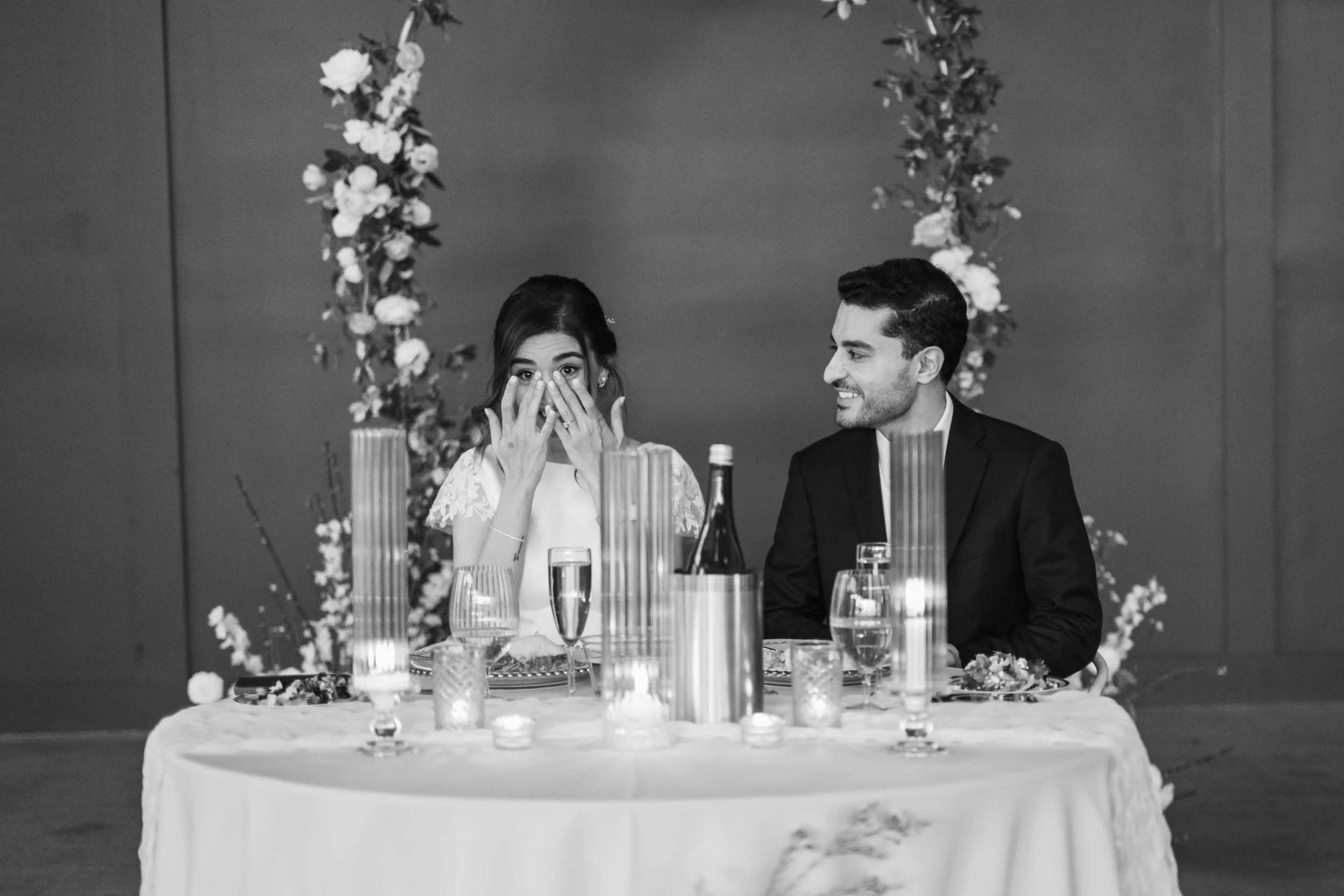 Black and white photo of a bride and groom sitting at a table at their wedding reception. Bride is wiping tears from her eyes