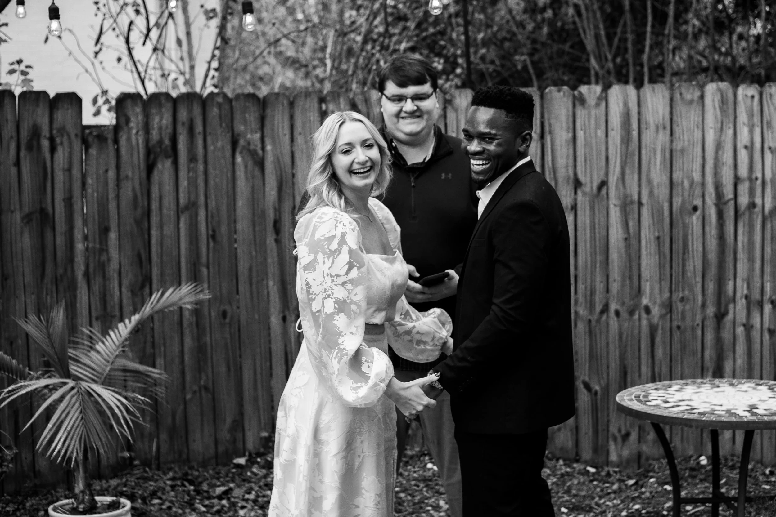 A black-and-white photo of a couple in wedding attire, holding hands and smiling, with a man and woman standing behind them. They are in a backyard with a wooden fence and string lights hanging above.
