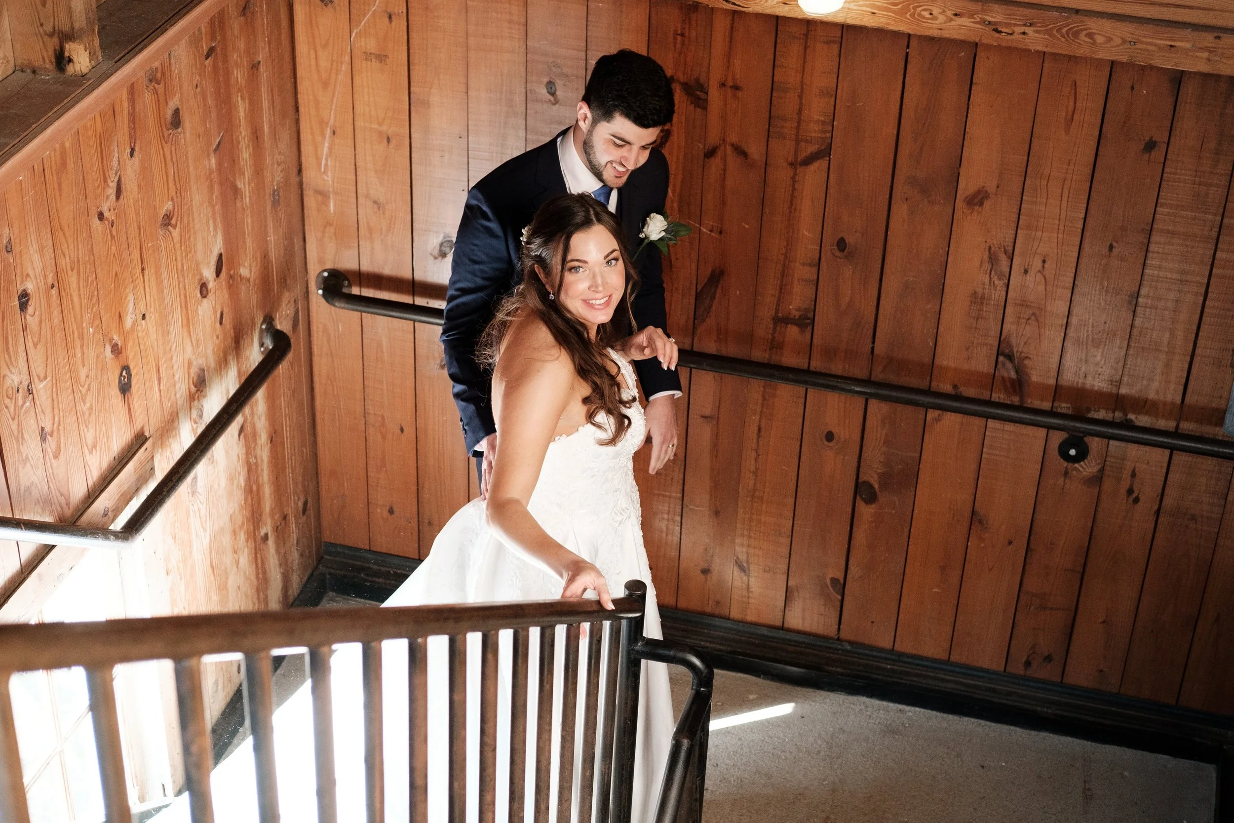 A bride in a white wedding gown taking a selfie with a groom in a black suit and white shirt behind her, inside a wooden staircase area at Founders Dairy Barn wedding venue